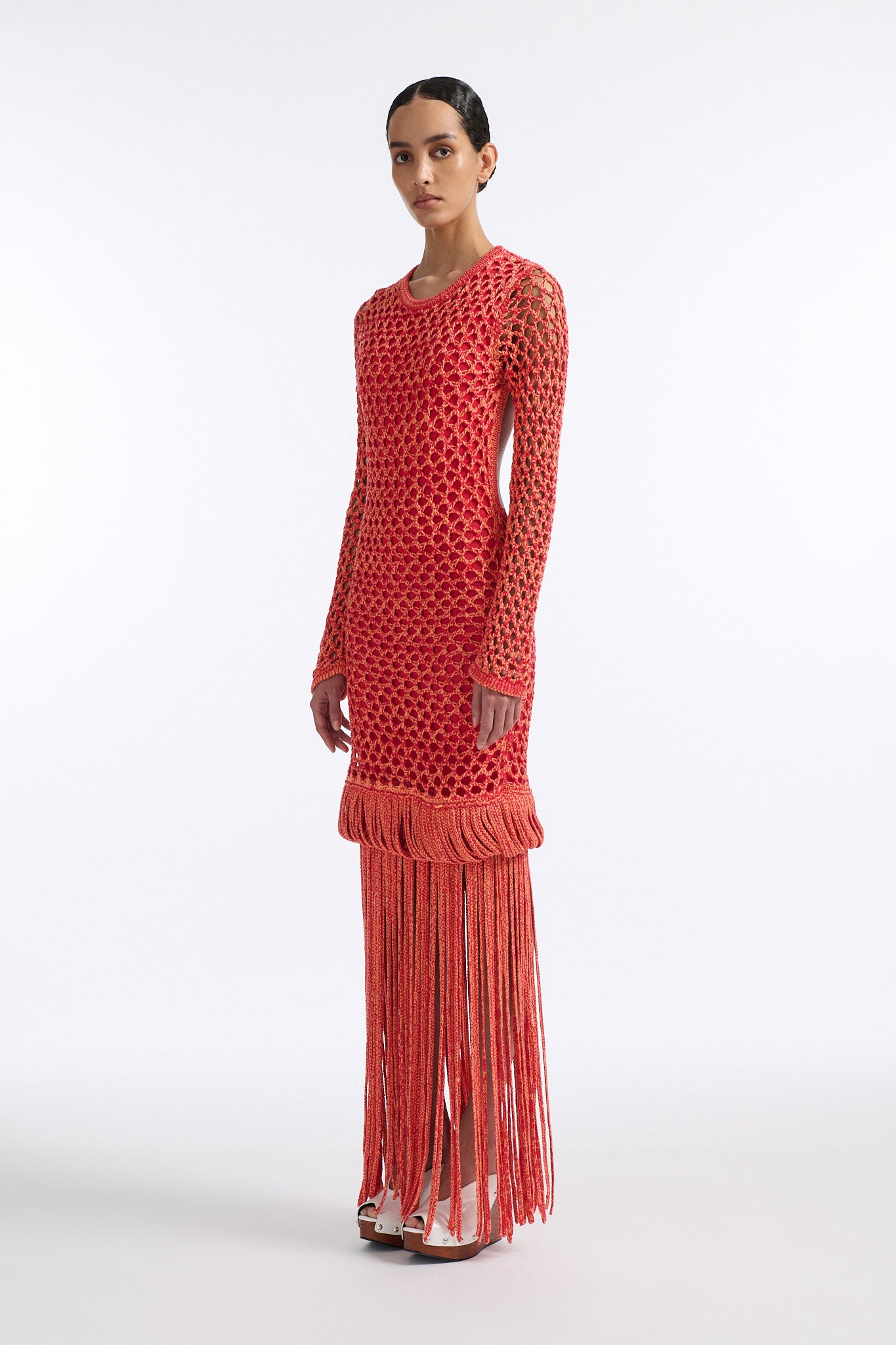 Female model with slicked back black hair in a bun wears red and orange mixed stitch chunky mesh long dress. Sleeves are open mesh and body is lined in red knit fabric. The hem of the dress has looped fringe that cascades down into floor length fringe.
