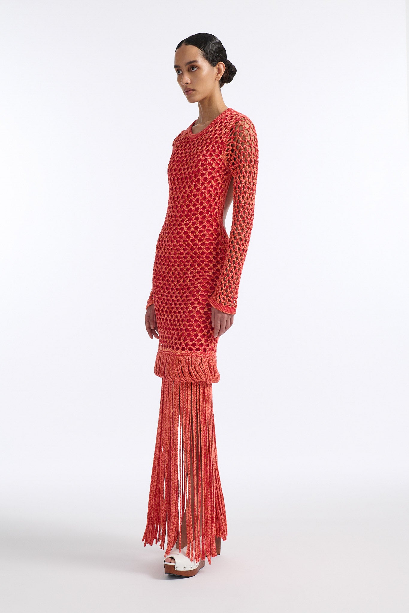 Female model with slicked back black hair in a bun wears red and orange mixed stitch chunky mesh long dress. Sleeves are open mesh and body is lined in red knit fabric. The hem of the dress has looped fringe that cascades down into floor length fringe.