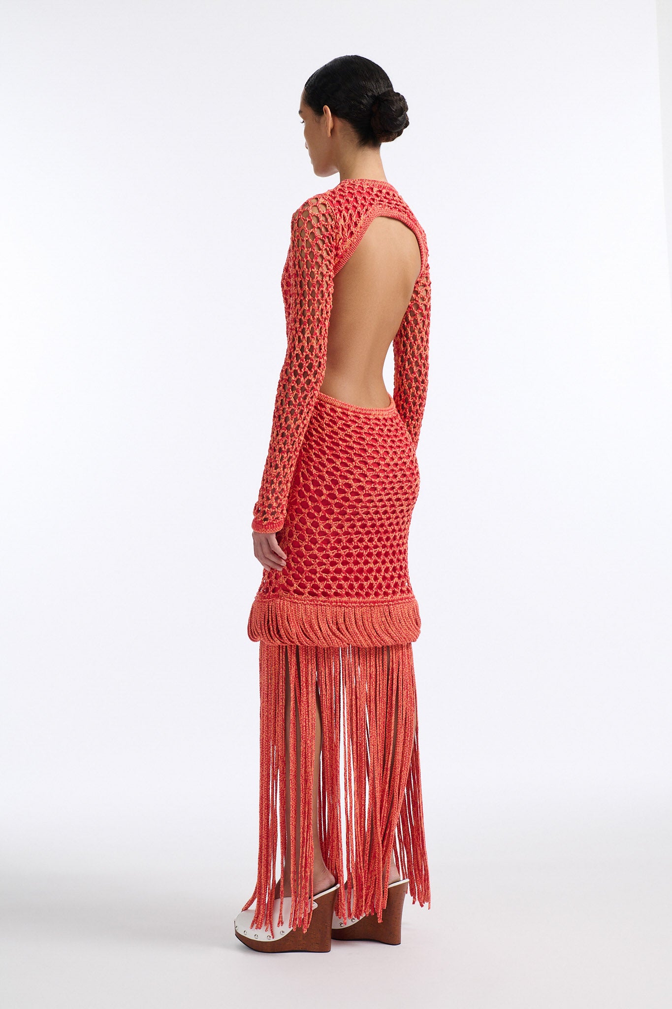Female model with slicked back black hair in a bun wears red and orange mixed stitch chunky mesh long dress. Sleeves are open mesh and body is lined in red knit fabric. The hem of the dress has looped fringe that cascades down into floor length fringe.