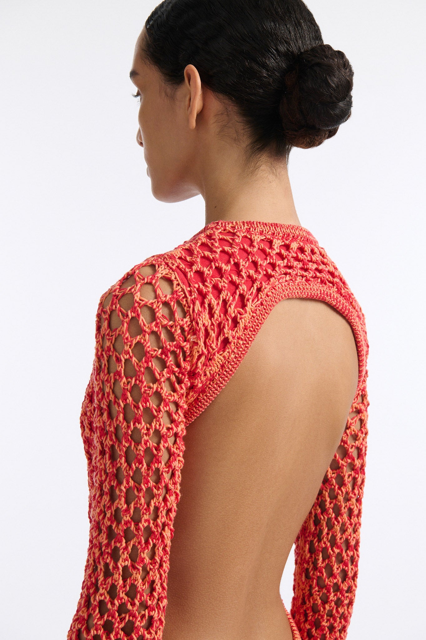 Female model with slicked back black hair in a bun wears red and orange mixed stitch chunky mesh long dress. Sleeves are open mesh and body is lined in red knit fabric. The hem of the dress has looped fringe that cascades down into floor length fringe.