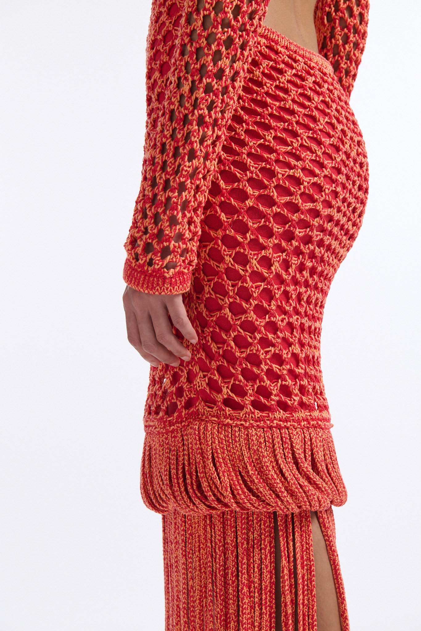 Female model with slicked back black hair in a bun wears red and orange mixed stitch chunky mesh long dress. Sleeves are open mesh and body is lined in red knit fabric. The hem of the dress has looped fringe that cascades down into floor length fringe.