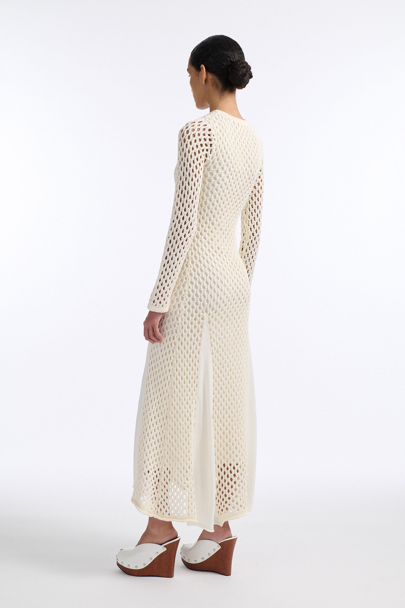 Female model with slicked back black hair in a bun wears white chunky mesh long dress. Dress has long sleeves that expose skin and a lining on the main body.