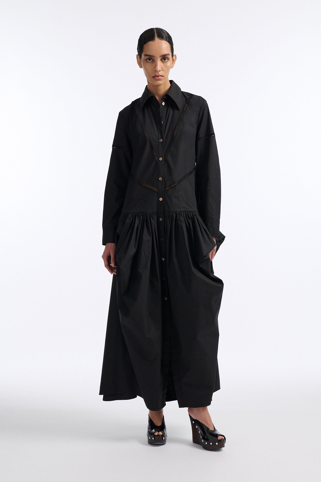 Female model with slicked back black hair in a bun wears black button down collared dress with diagonal hand stitched detailing along front, back and sleeves. The waist of the skirt is rouched to provide volume. The model also wears white palm cutout denim.
