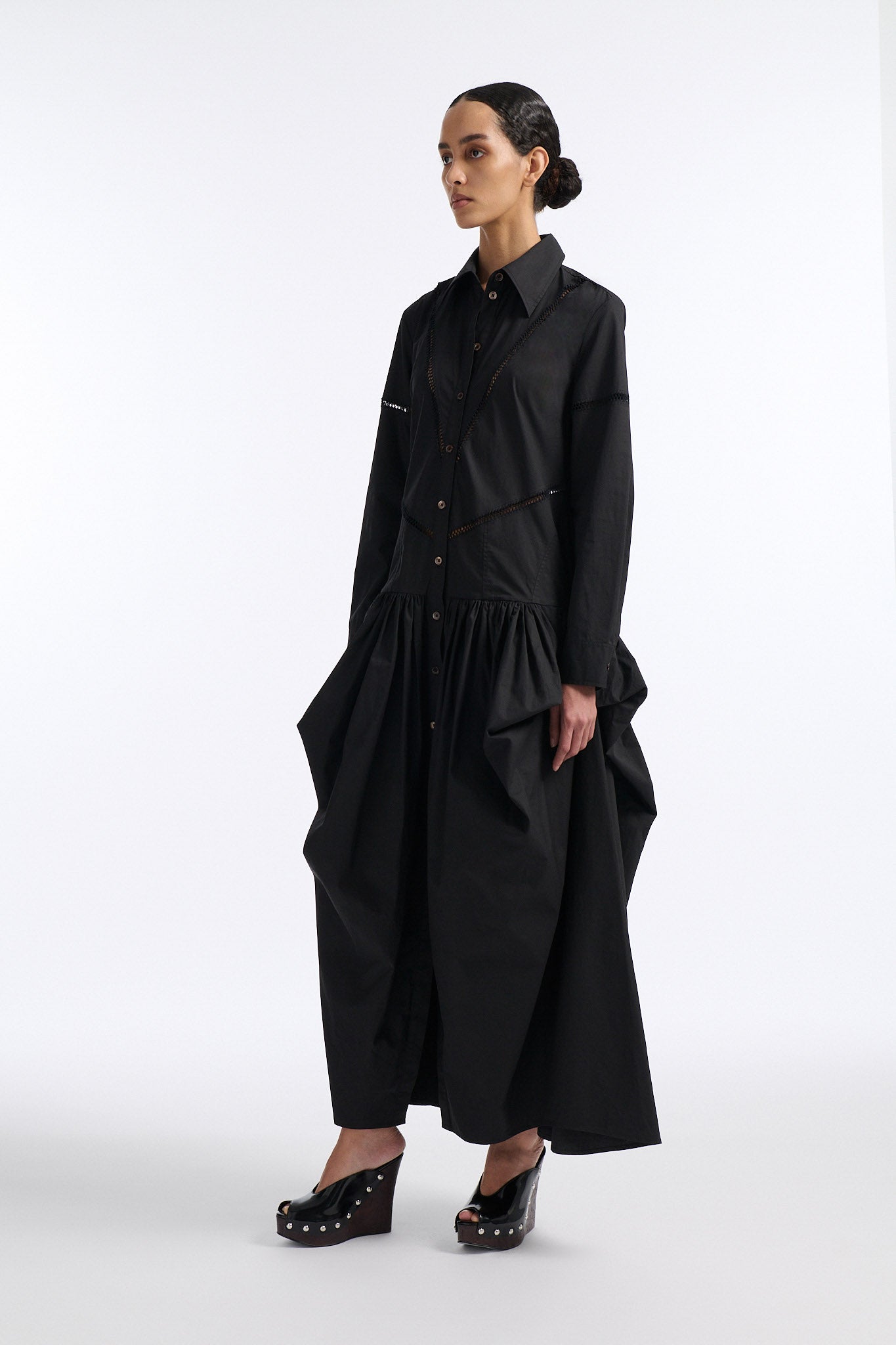 Female model with slicked back black hair in a bun wears black button down collared dress with diagonal hand stitched detailing along front, back and sleeves. The waist of the skirt is rouched to provide volume. The model also wears white palm cutout denim.