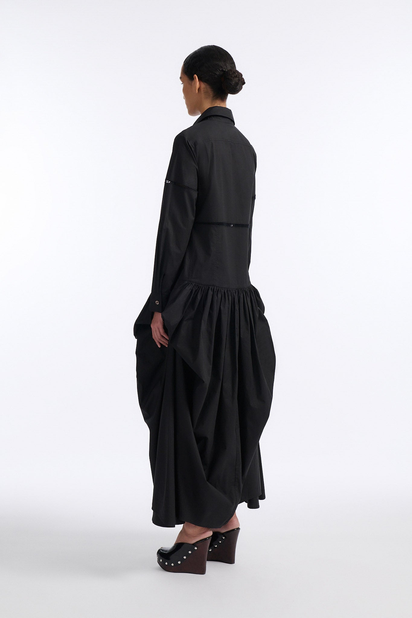 Female model with slicked back black hair in a bun wears black button down collared dress with diagonal hand stitched detailing along front, back and sleeves. The waist of the skirt is rouched to provide volume. The model also wears white palm cutout denim.