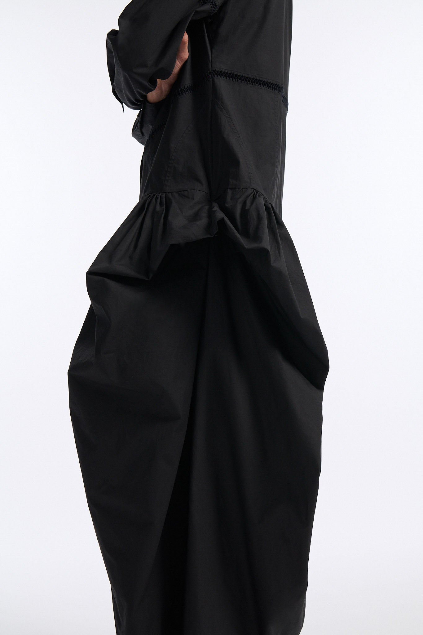 Female model with slicked back black hair in a bun wears black button down collared dress with diagonal hand stitched detailing along front, back and sleeves. The waist of the skirt is rouched to provide volume. The model also wears white palm cutout denim.