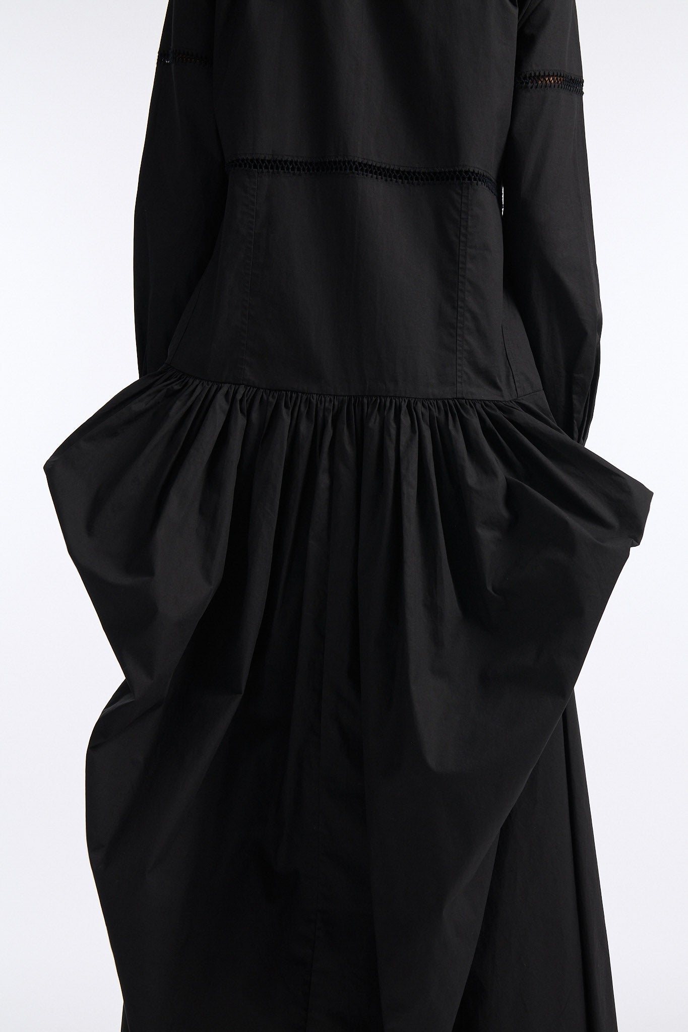 Female model with slicked back black hair in a bun wears black button down collared dress with diagonal hand stitched detailing along front, back and sleeves. The waist of the skirt is rouched to provide volume. The model also wears white palm cutout denim.