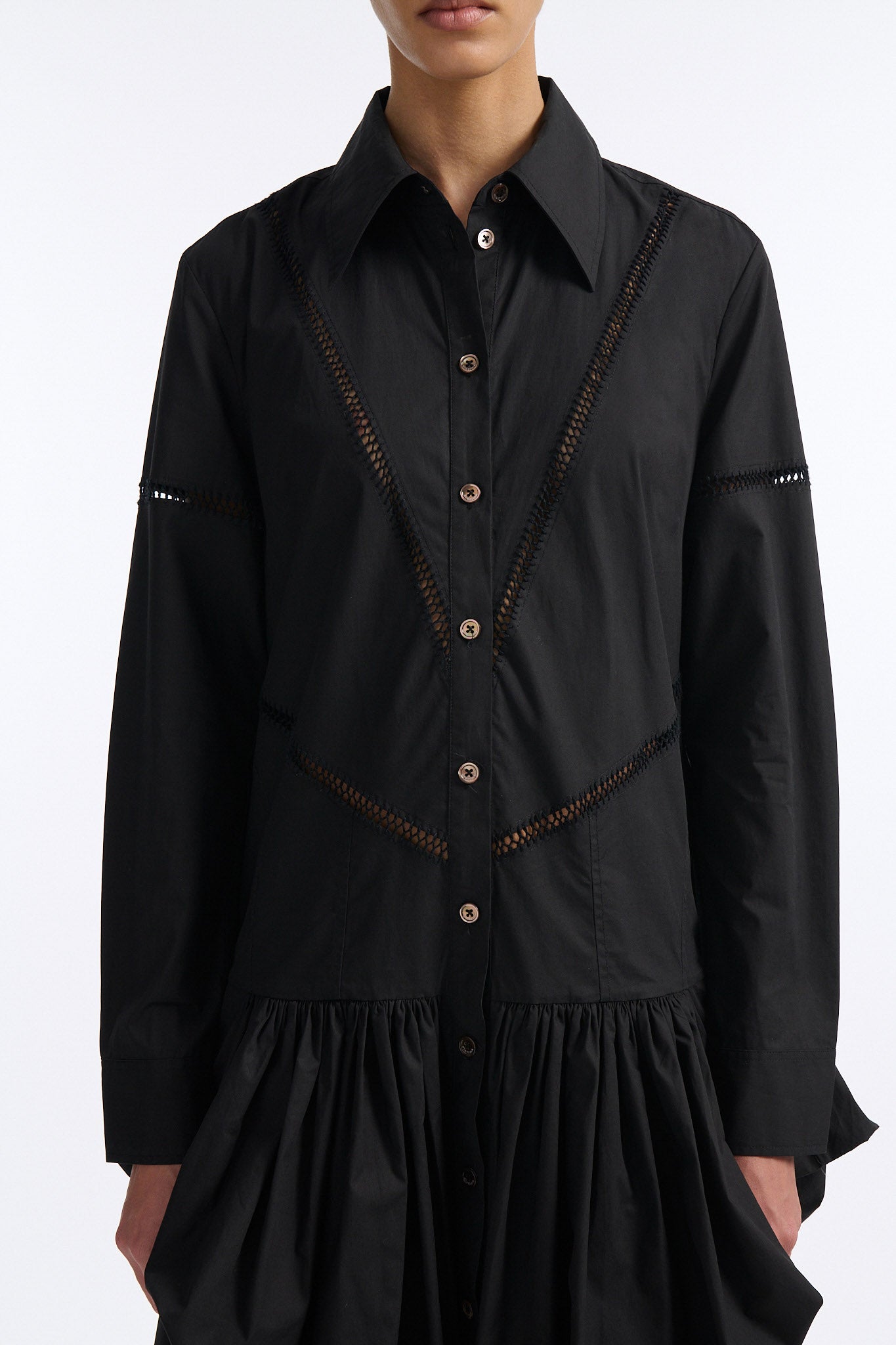 Female model with slicked back black hair in a bun wears black button down collared dress with diagonal hand stitched detailing along front, back and sleeves. The waist of the skirt is rouched to provide volume. The model also wears white palm cutout denim.