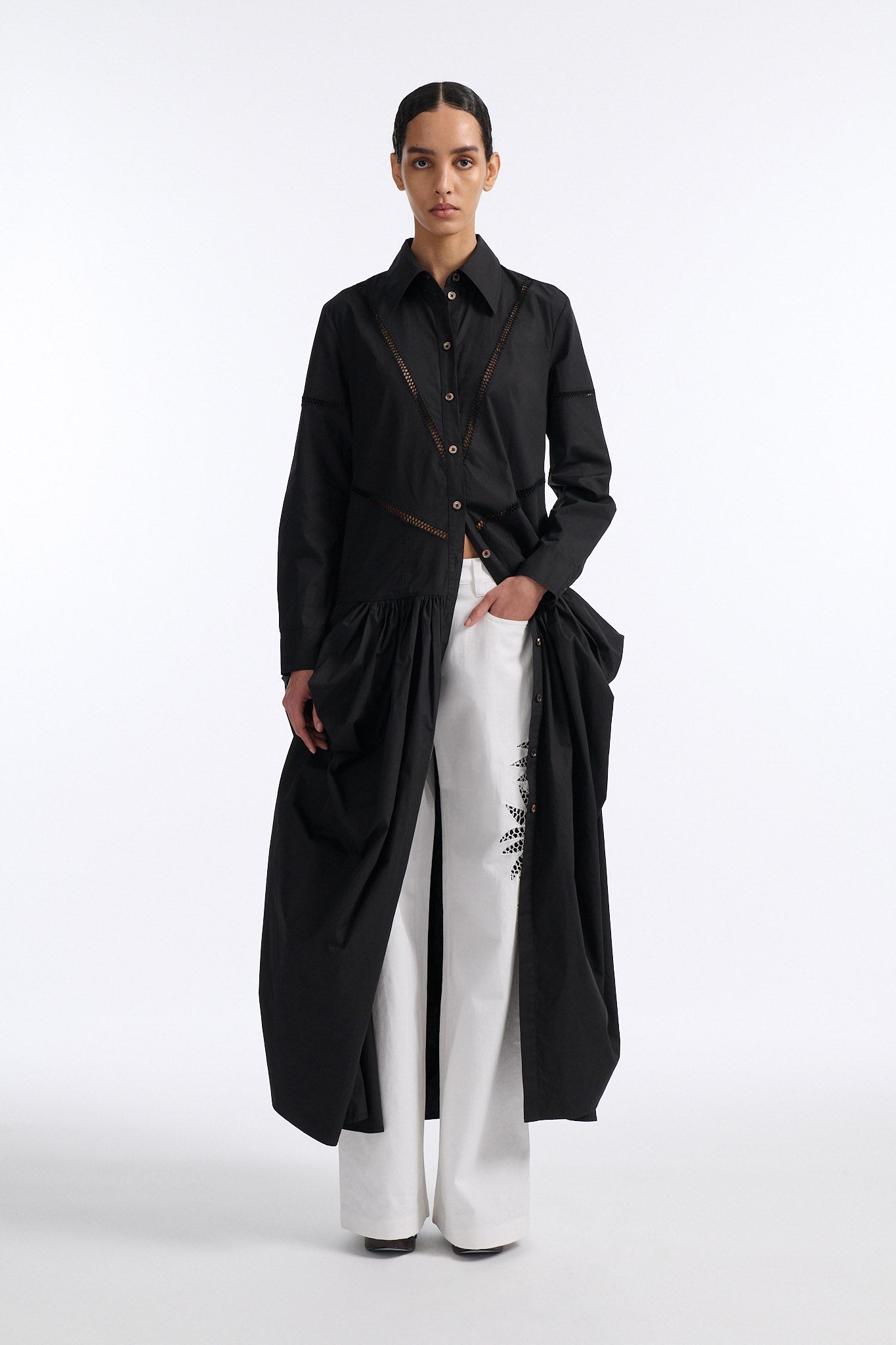 Female model with slicked back black hair in a bun wears black button down collared dress with diagonal hand stitched detailing along front, back and sleeves. The waist of the skirt is rouched to provide volume. The model also wears white palm cutout denim.