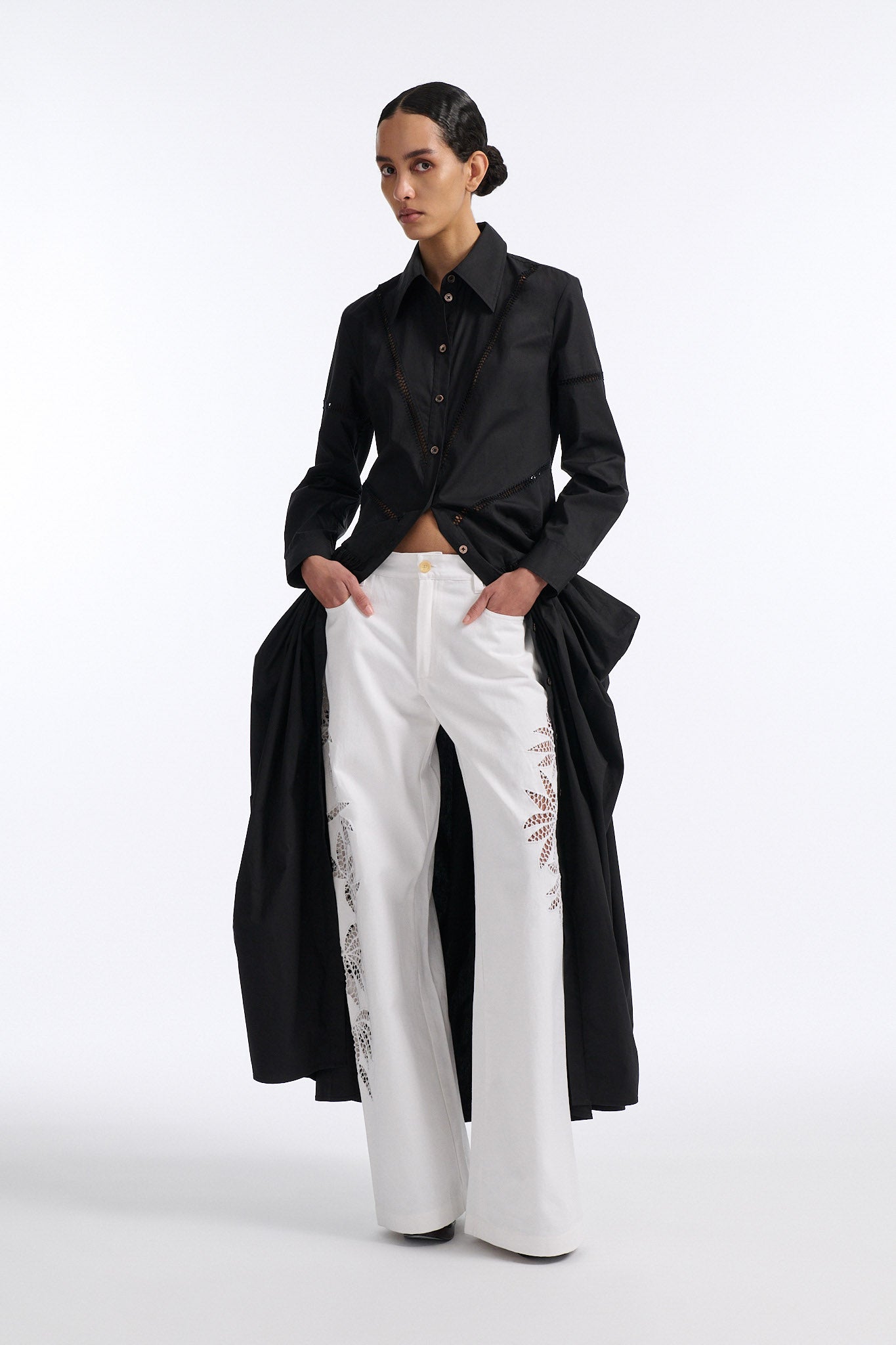 Female model with slicked back black hair in a bun wears black button down collared dress with diagonal hand stitched detailing along front, back and sleeves. The waist of the skirt is rouched to provide volume. The model also wears white palm cutout denim.