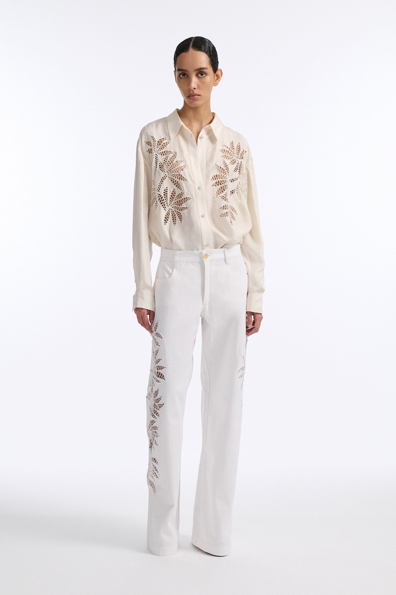 Female model with slicked back black hair in a bun wears white mid-waist denim jeans with palm leaf cutouts on either side. Model also wears matching palm leaf cutout silk button down.