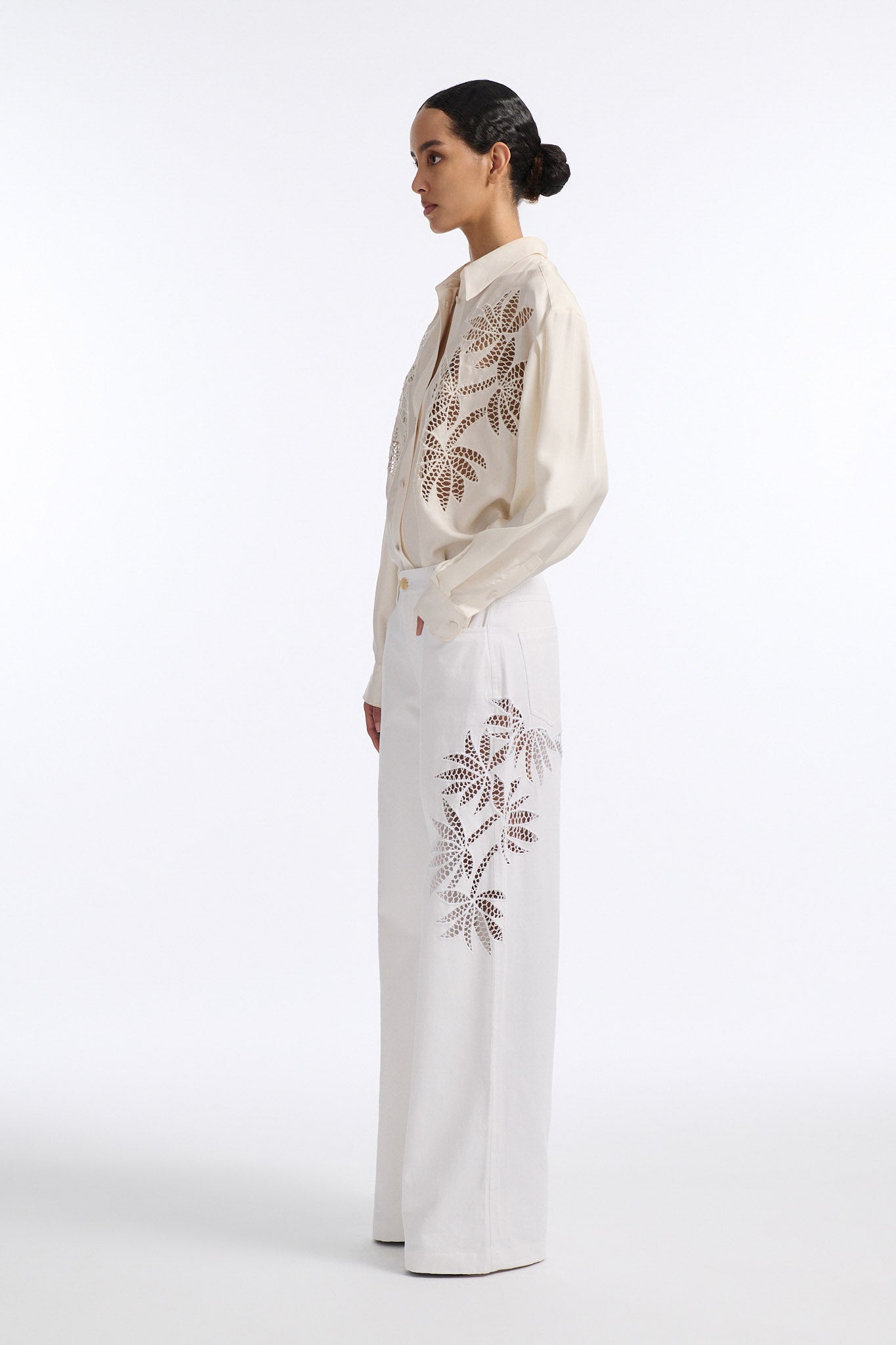 Female model with slicked back black hair in a bun wears white mid-waist denim jeans with palm leaf cutouts on either side. Model also wears matching palm leaf cutout silk button down.