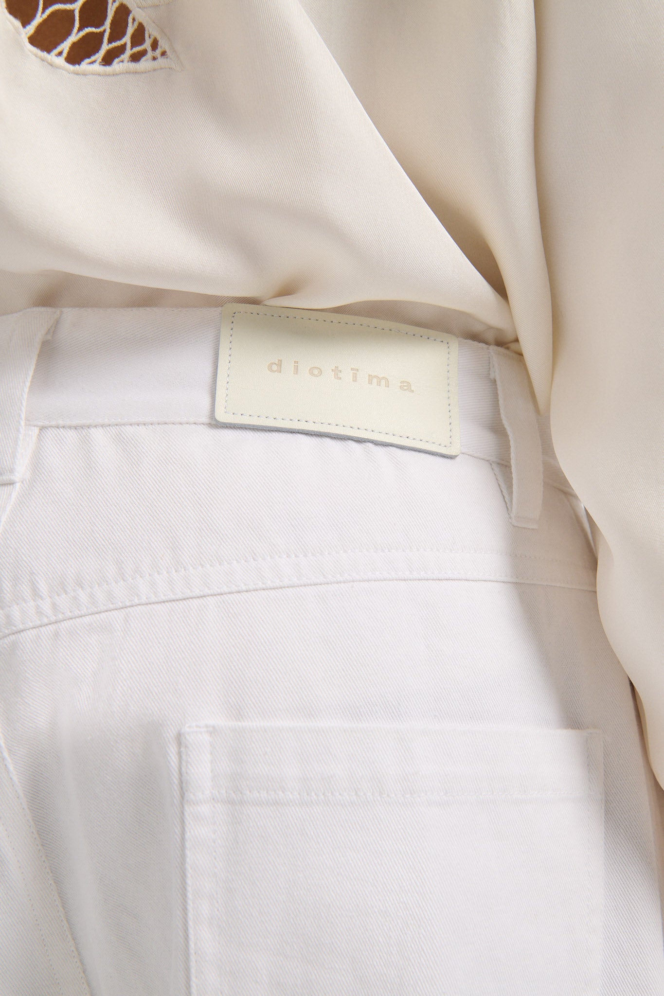 Female model with slicked back black hair in a bun wears white mid-waist denim jeans with palm leaf cutouts on either side. Model also wears matching palm leaf cutout silk button down.