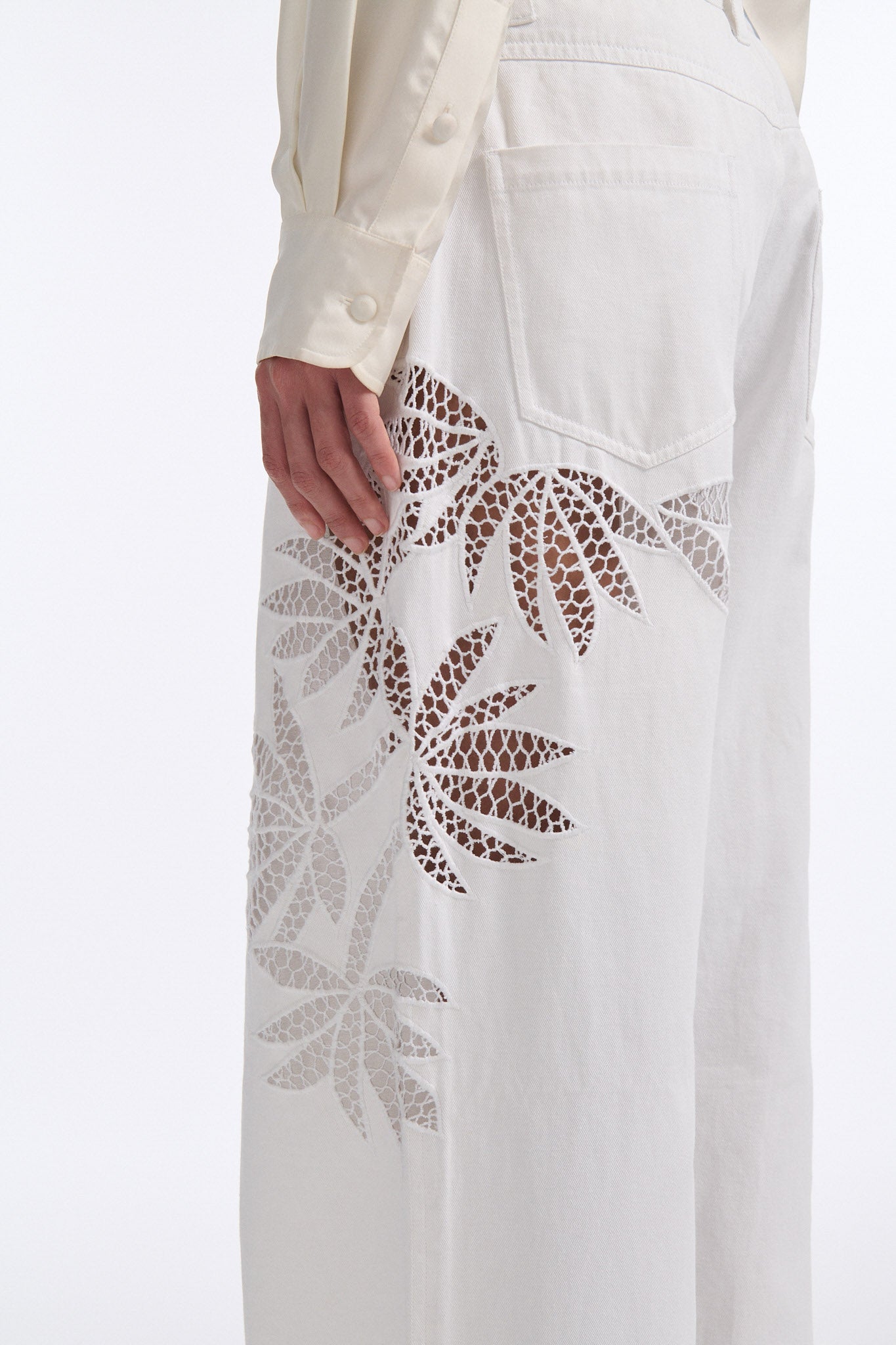 Female model with slicked back black hair in a bun wears white mid-waist denim jeans with palm leaf cutouts on either side. Model also wears matching palm leaf cutout silk button down.