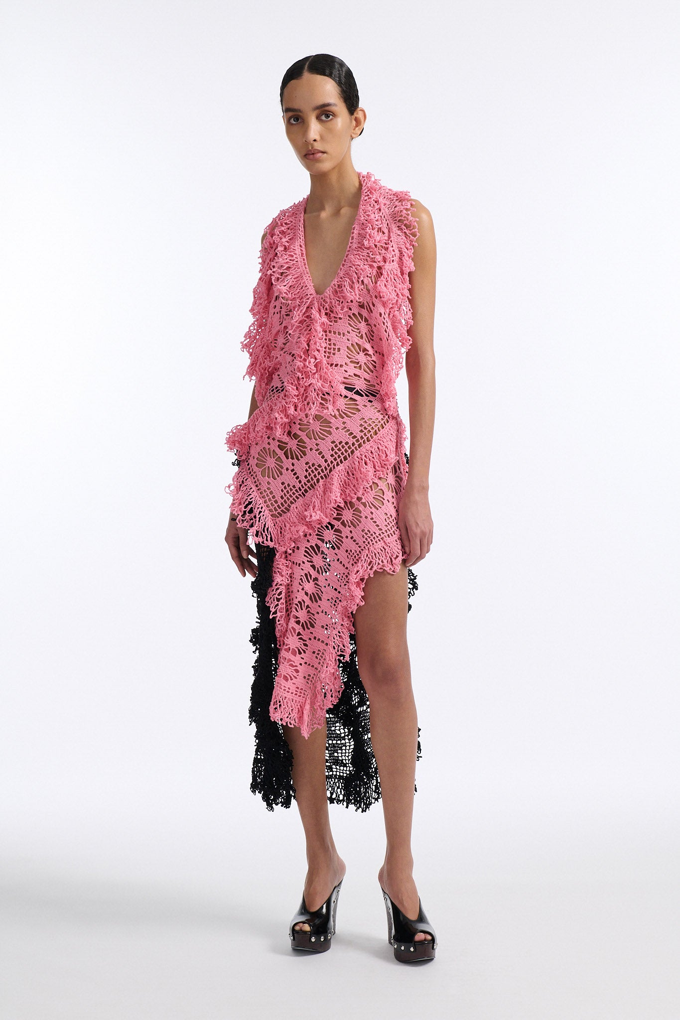 Female model with slicked back black hair in a bun wears V-neck crochet panel dress. The entire dress is made of flat crochet panels with fringe hems, the front of the dress is ginger pink and the back is open back in black crochet.