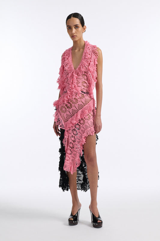 Female model with slicked back black hair in a bun wears V-neck crochet panel dress. The entire dress is made of flat crochet panels with fringe hems, the front of the dress is ginger pink and the back is open back in black crochet.