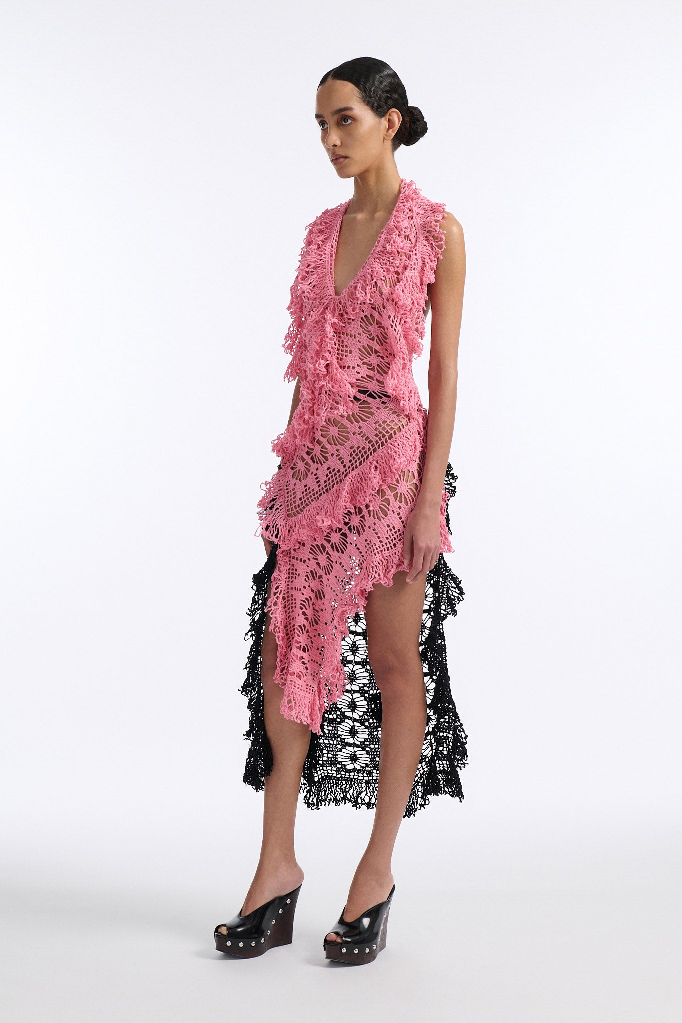Female model with slicked back black hair in a bun wears V-neck crochet panel dress. The entire dress is made of flat crochet panels with fringe hems, the front of the dress is ginger pink and the back is open back in black crochet.