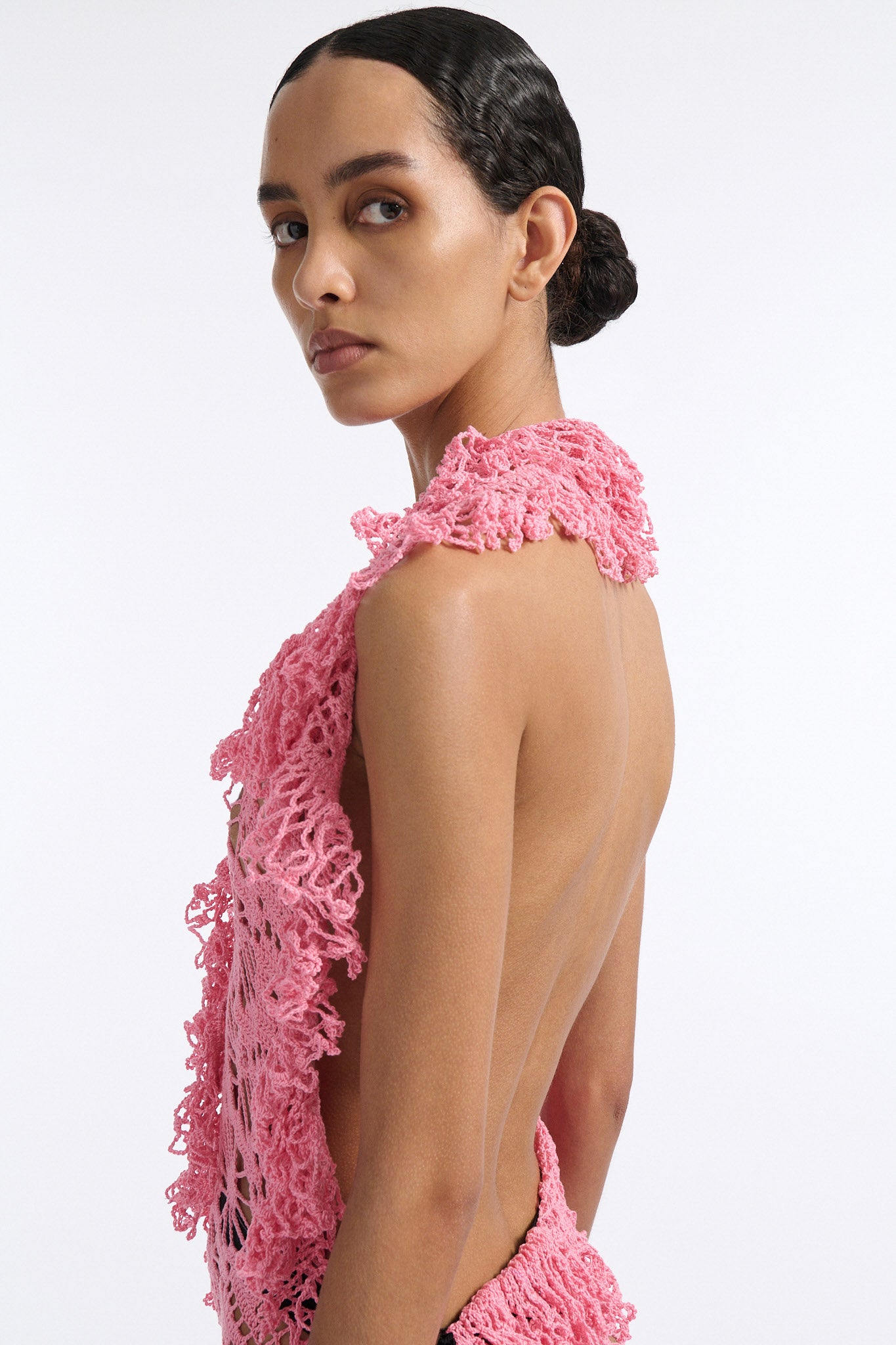 Female model with slicked back black hair in a bun wears V-neck crochet panel dress. The entire dress is made of flat crochet panels with fringe hems, the front of the dress is ginger pink and the back is open back in black crochet.