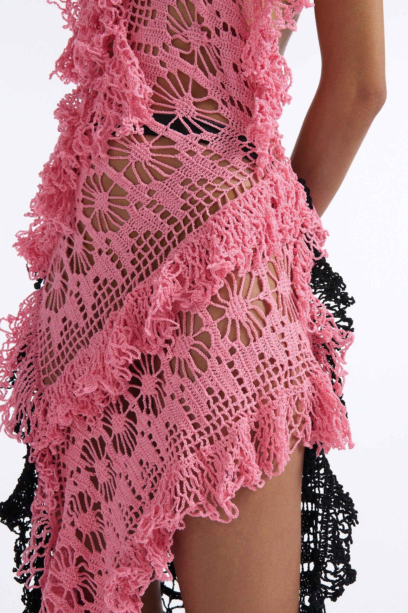 Female model with slicked back black hair in a bun wears V-neck crochet panel dress. The entire dress is made of flat crochet panels with fringe hems, the front of the dress is ginger pink and the back is open back in black crochet.