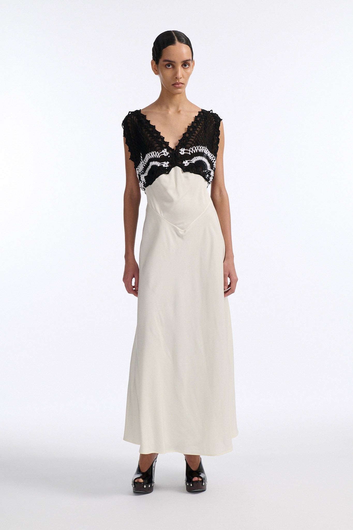 Female model with slicked back black hair in a bun wears long white shift dress with triangle crochet paneling on check. Crochet is black with two white stripes and is attacked to two straps that criss-cross over an open back.