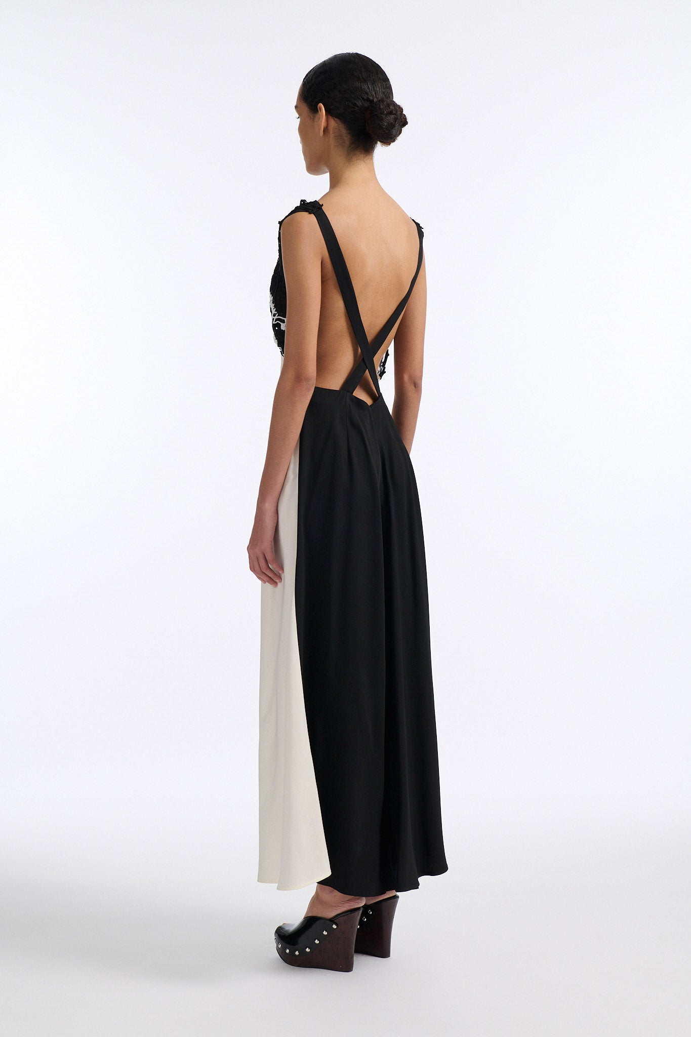 Female model with slicked back black hair in a bun wears long white shift dress with triangle crochet paneling on check. Crochet is black with two white stripes and is attacked to two straps that criss-cross over an open back.