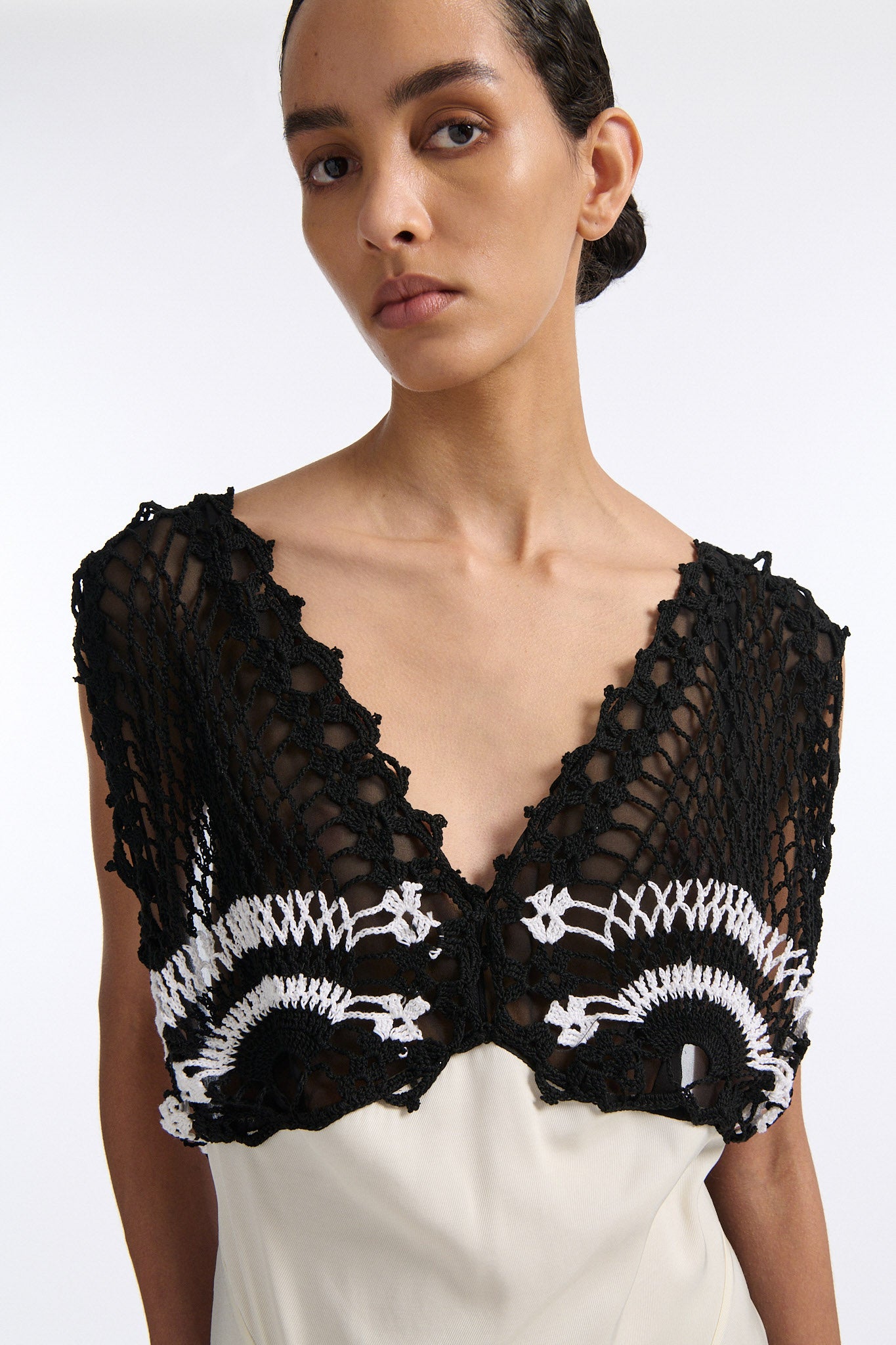 Female model with slicked back black hair in a bun wears long white shift dress with triangle crochet paneling on check. Crochet is black with two white stripes and is attacked to two straps that criss-cross over an open back.