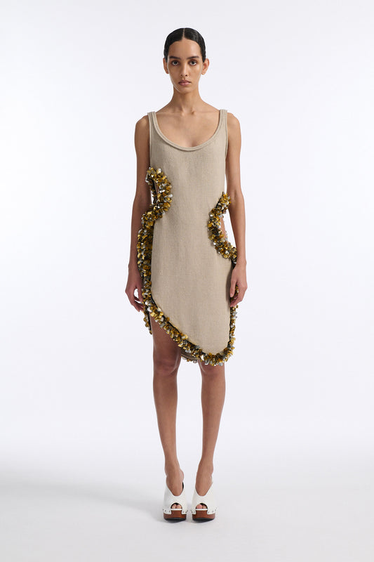 Female model with slicked back black hair in a bun wears beige linen tank dress with curved hem and asymmetric cutouts on either side. Hem and cutouts are lined with melted metallic embellishments in gold and silver to provide an oyster illusion.