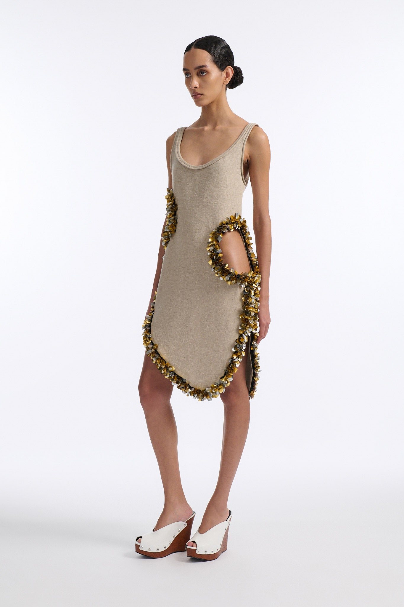 Female model with slicked back black hair in a bun wears beige linen tank dress with curved hem and asymmetric cutouts on either side. Hem and cutouts are lined with melted metallic embellishments in gold and silver to provide an oyster illusion.