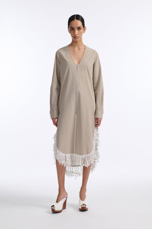 Female model with slicked back black hair in a bun wears long beige V-neck cotton dress with curved hem and slit on both sides. Hem has white beaded fringe and the front of the dress has a long vertical stripe of hand stitch detailing.