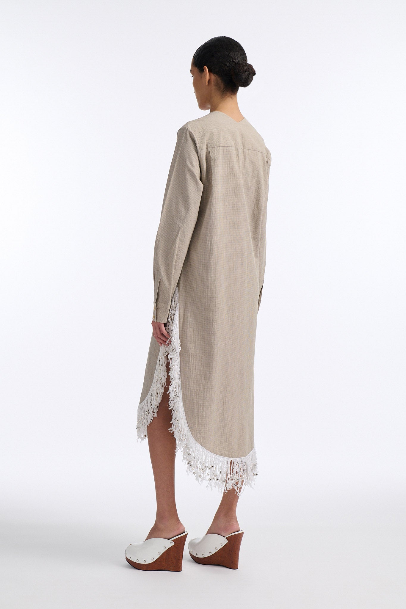 Female model with slicked back black hair in a bun wears long beige V-neck cotton dress with curved hem and slit on both sides. Hem has white beaded fringe and the front of the dress has a long vertical stripe of hand stitch detailing.