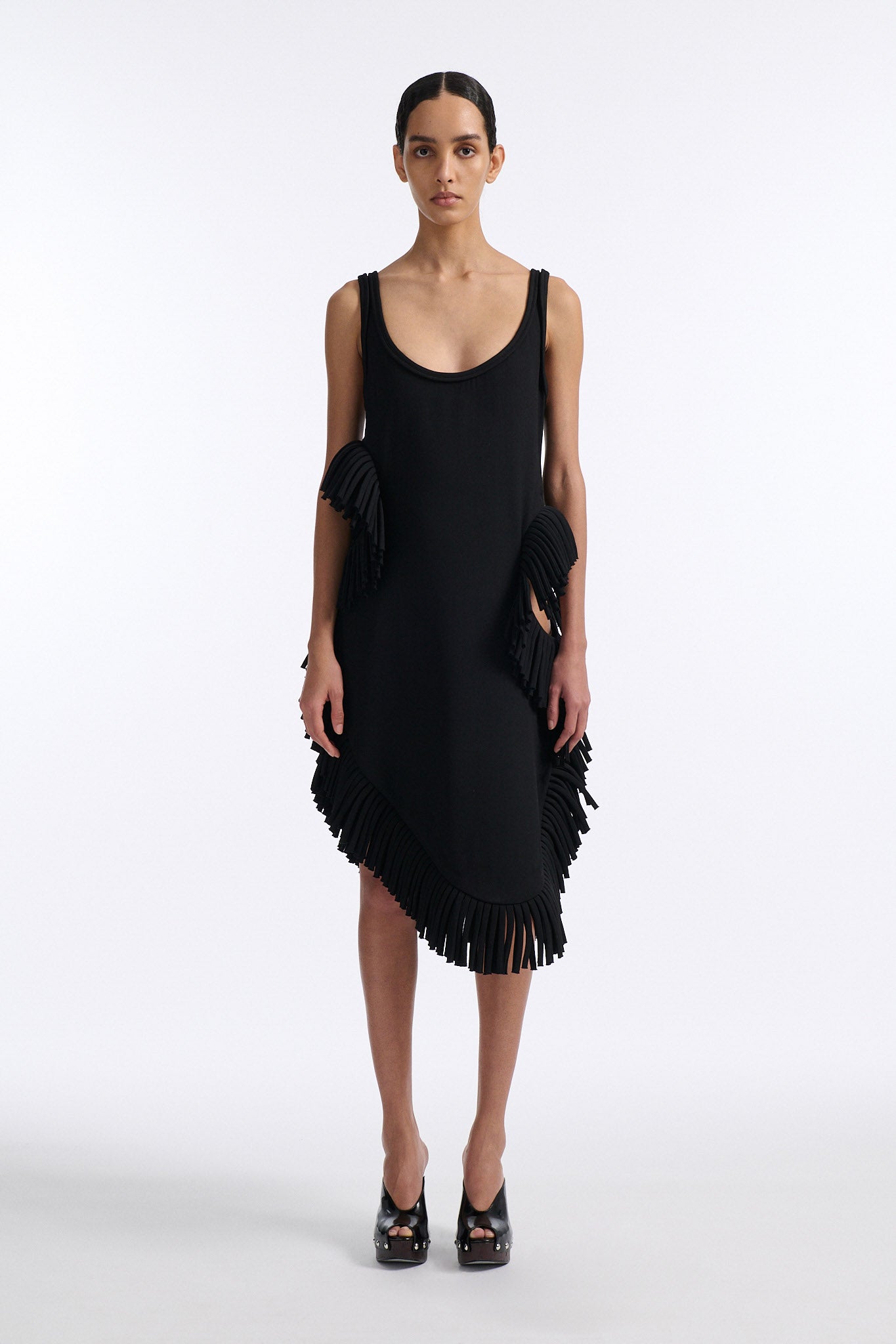 Female model with slicked back black hair in a bun wears midi black fine wool tank dress with curved hem and asymmetric side cutouts. There is rolled black fringe at side cutouts and along bottom hem.
