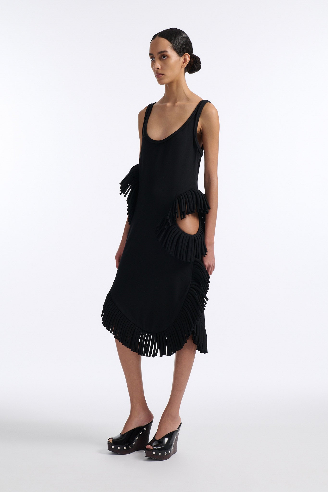 Female model with slicked back black hair in a bun wears midi black fine wool tank dress with curved hem and asymmetric side cutouts. There is rolled black fringe at side cutouts and along bottom hem.
