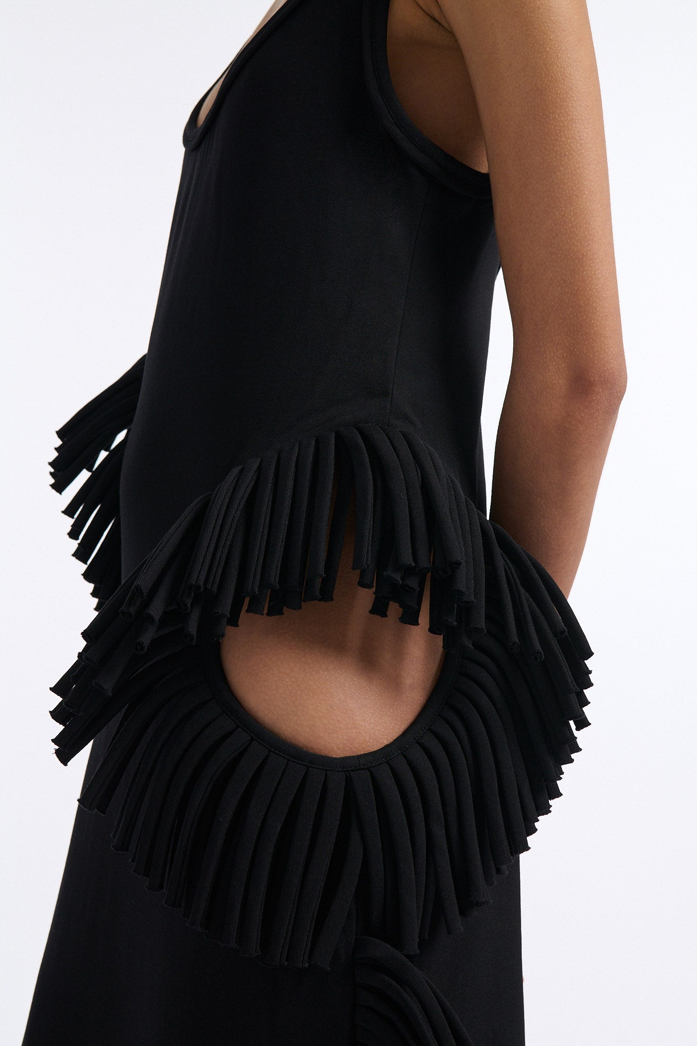 Female model with slicked back black hair in a bun wears midi black fine wool tank dress with curved hem and asymmetric side cutouts. There is rolled black fringe at side cutouts and along bottom hem.