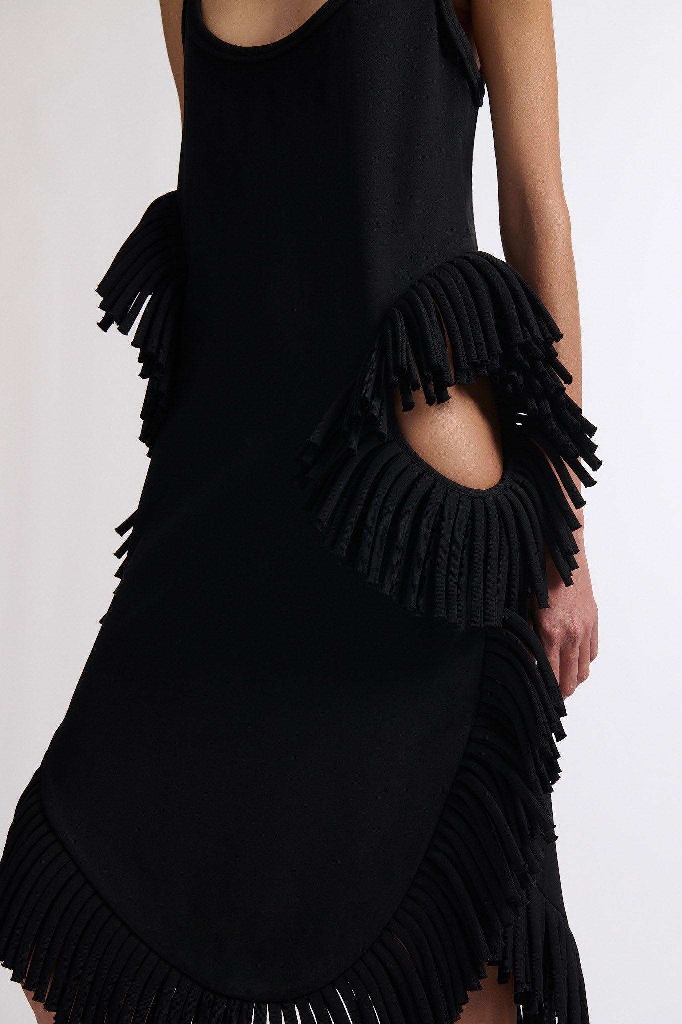 Female model with slicked back black hair in a bun wears midi black fine wool tank dress with curved hem and asymmetric side cutouts. There is rolled black fringe at side cutouts and along bottom hem.
