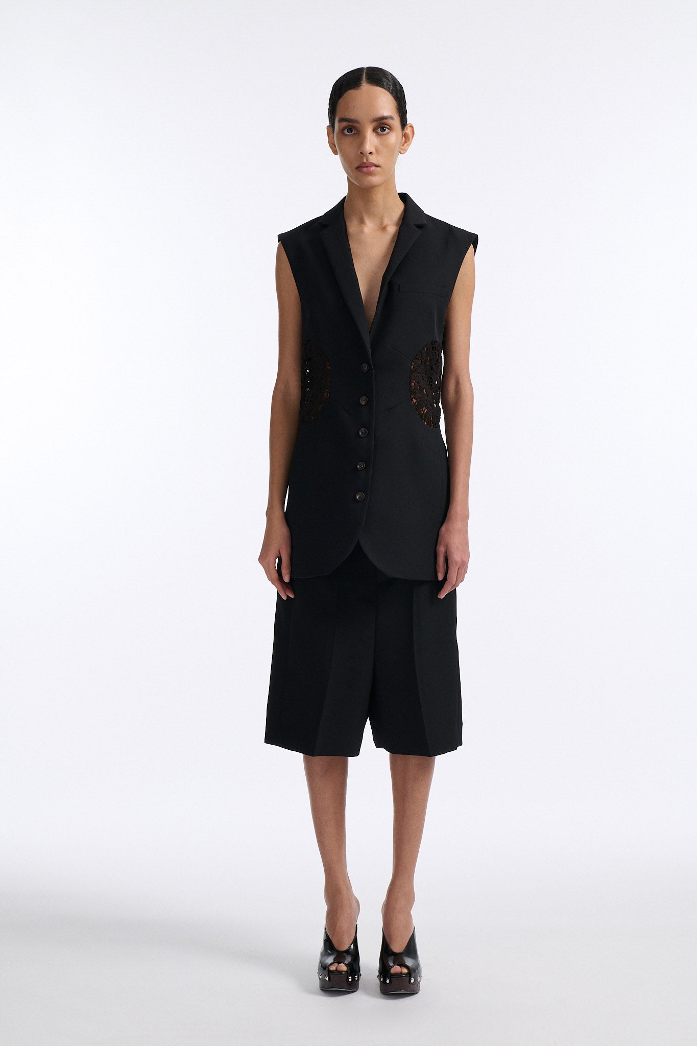 Female model with slicked back black hair in a bun wears fine black wool 5 button vest with black crochet circle inserts on either side. Model also wears matching pleated black wool shorts and black wedges.
