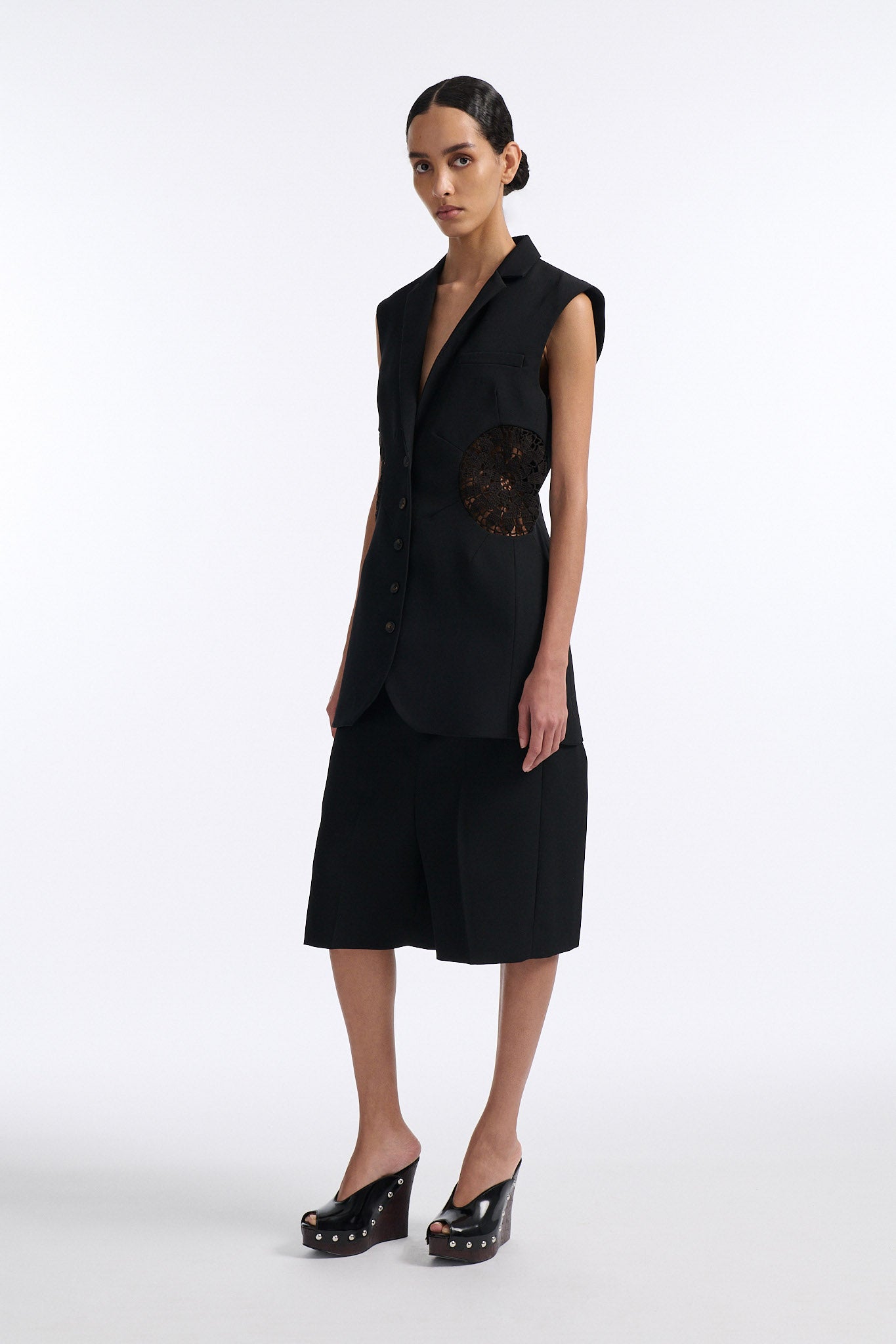 Female model with slicked back black hair in a bun wears fine black wool 5 button vest with black crochet circle inserts on either side. Model also wears matching pleated black wool shorts and black wedges.