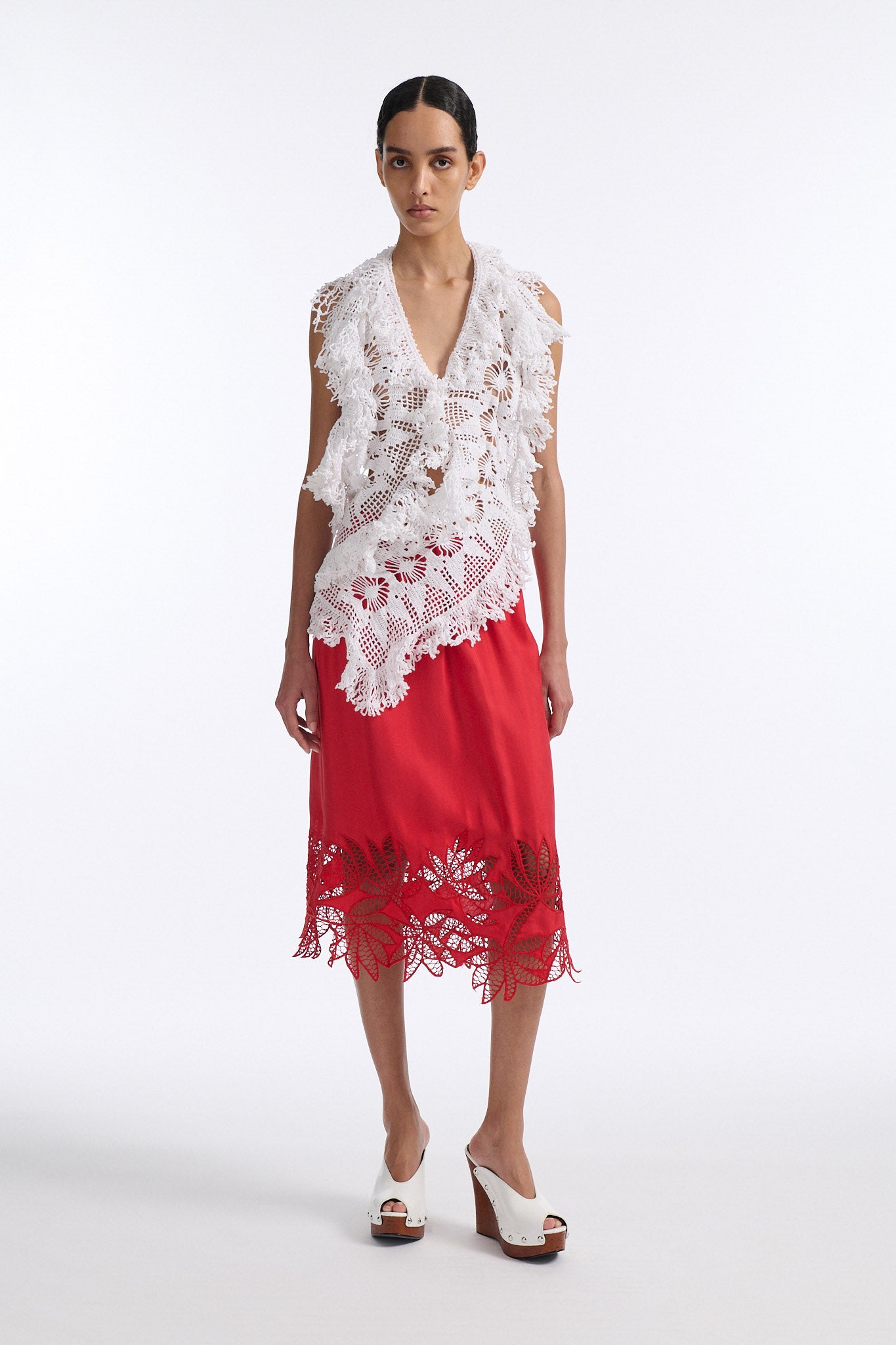 Female model with slicked back black hair in a bun wears white crochet V-neck sleeveless top with fringe crochet finishings and open back. Model also wears red silk shift skirt with cutout palm leaf hem and elastic waist.