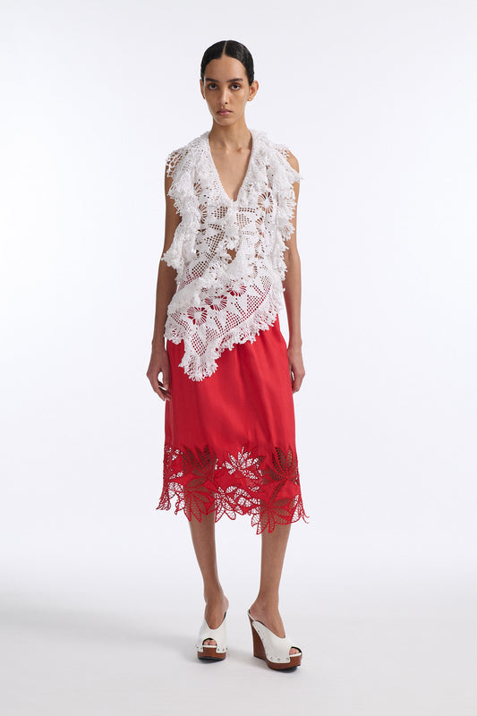 Female model with slicked back black hair in a bun wears white crochet V-neck sleeveless top with fringe crochet finishings and open back. Model also wears red silk shift skirt with cutout palm leaf hem and elastic waist.