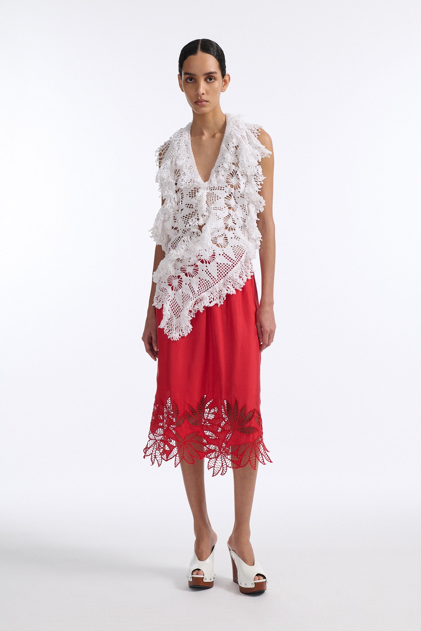 Female model with slicked back black hair in a bun wears white crochet V-neck sleeveless top with fringe crochet finishings and open back. Model also wears red silk shift skirt with cutout palm leaf hem and elastic waist.