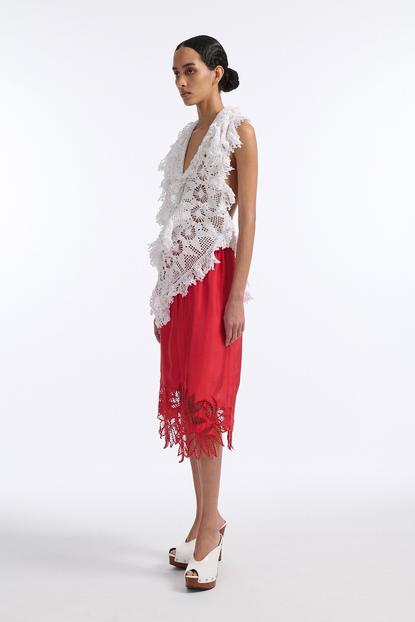 Female model with slicked back black hair in a bun wears white crochet V-neck sleeveless top with fringe crochet finishings and open back. Model also wears red silk shift skirt with cutout palm leaf hem and elastic waist.