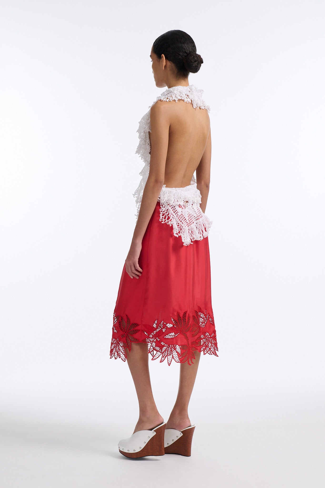 Female model with slicked back black hair in a bun wears white crochet V-neck sleeveless top with fringe crochet finishings and open back. Model also wears red silk shift skirt with cutout palm leaf hem and elastic waist.