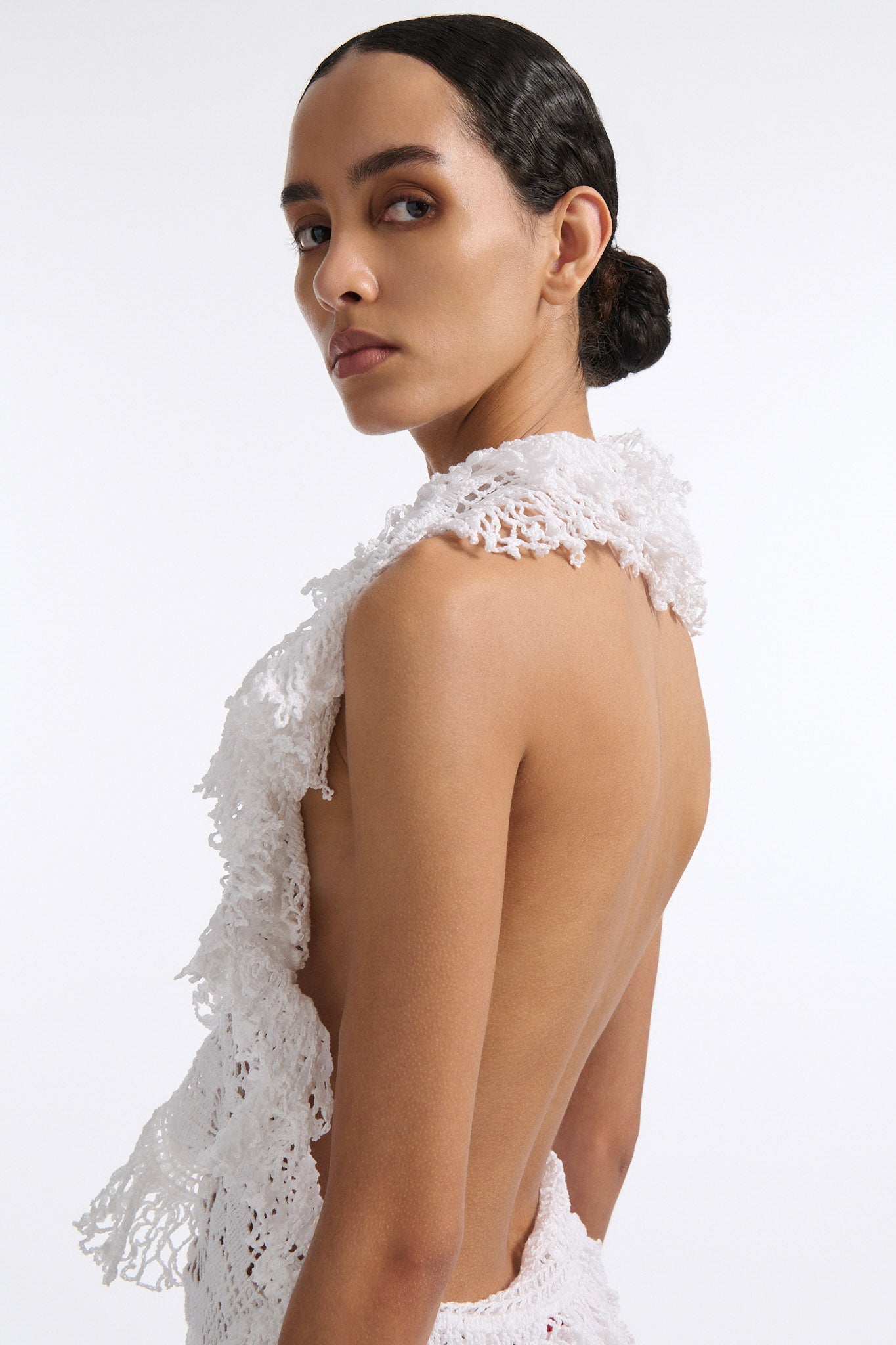 Female model with slicked back black hair in a bun wears white crochet V-neck sleeveless top with fringe crochet finishings and open back. Model also wears red silk shift skirt with cutout palm leaf hem and elastic waist.