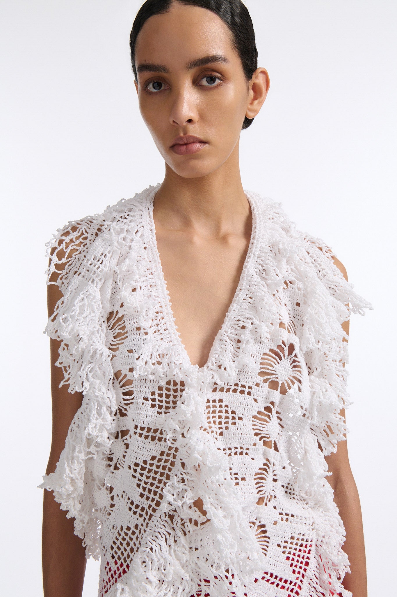 Female model with slicked back black hair in a bun wears white crochet V-neck sleeveless top with fringe crochet finishings and open back. Model also wears red silk shift skirt with cutout palm leaf hem and elastic waist.