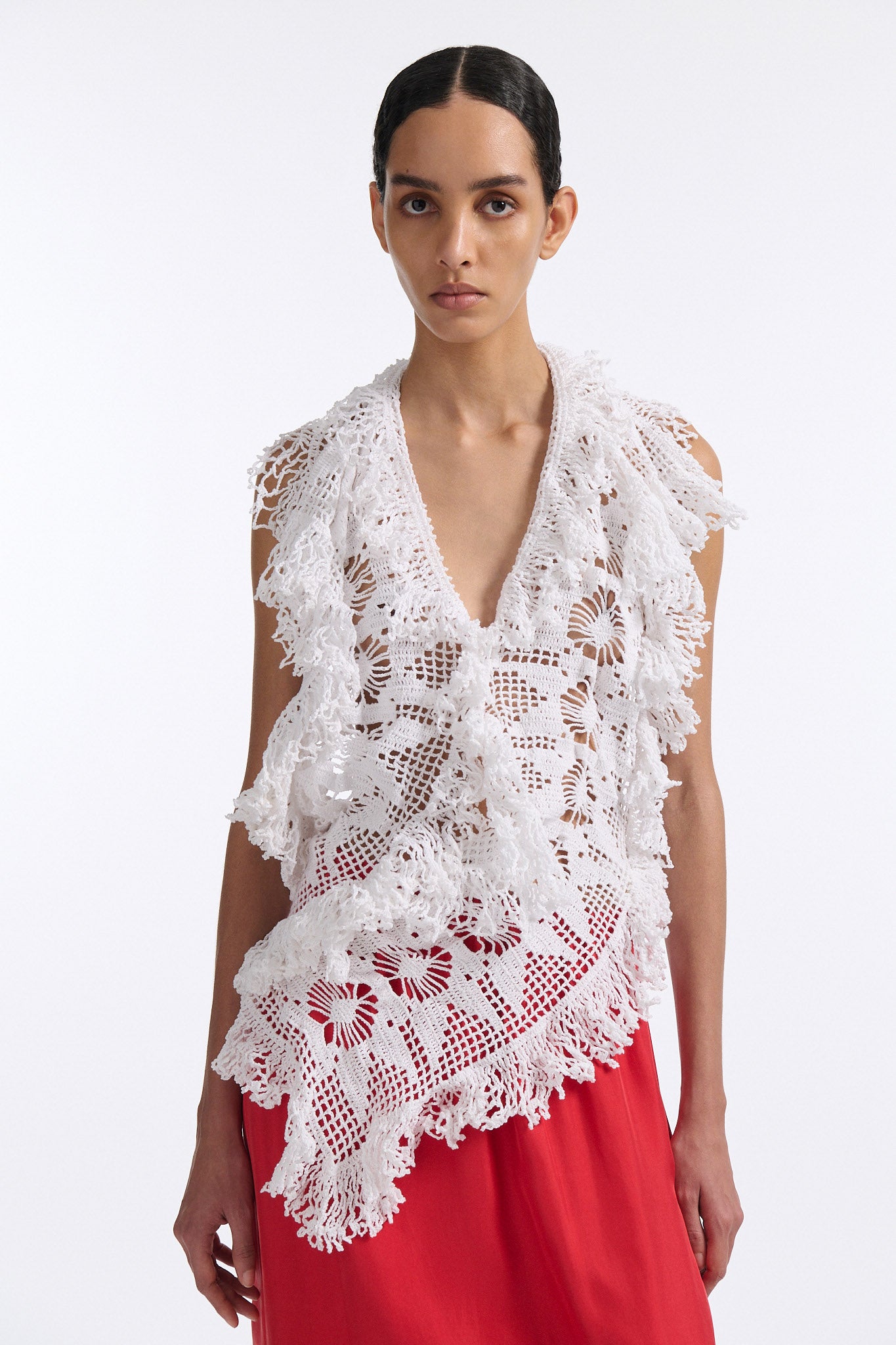 Female model with slicked back black hair in a bun wears white crochet V-neck sleeveless top with fringe crochet finishings and open back. Model also wears red silk shift skirt with cutout palm leaf hem and elastic waist.
