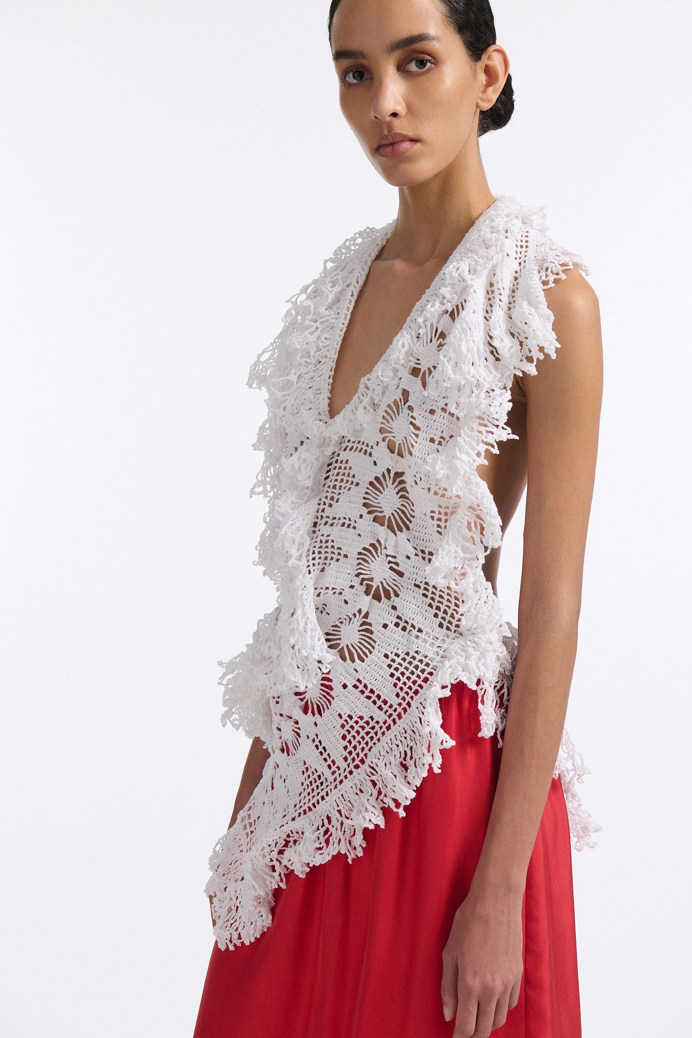 Female model with slicked back black hair in a bun wears white crochet V-neck sleeveless top with fringe crochet finishings and open back. Model also wears red silk shift skirt with cutout palm leaf hem and elastic waist.
