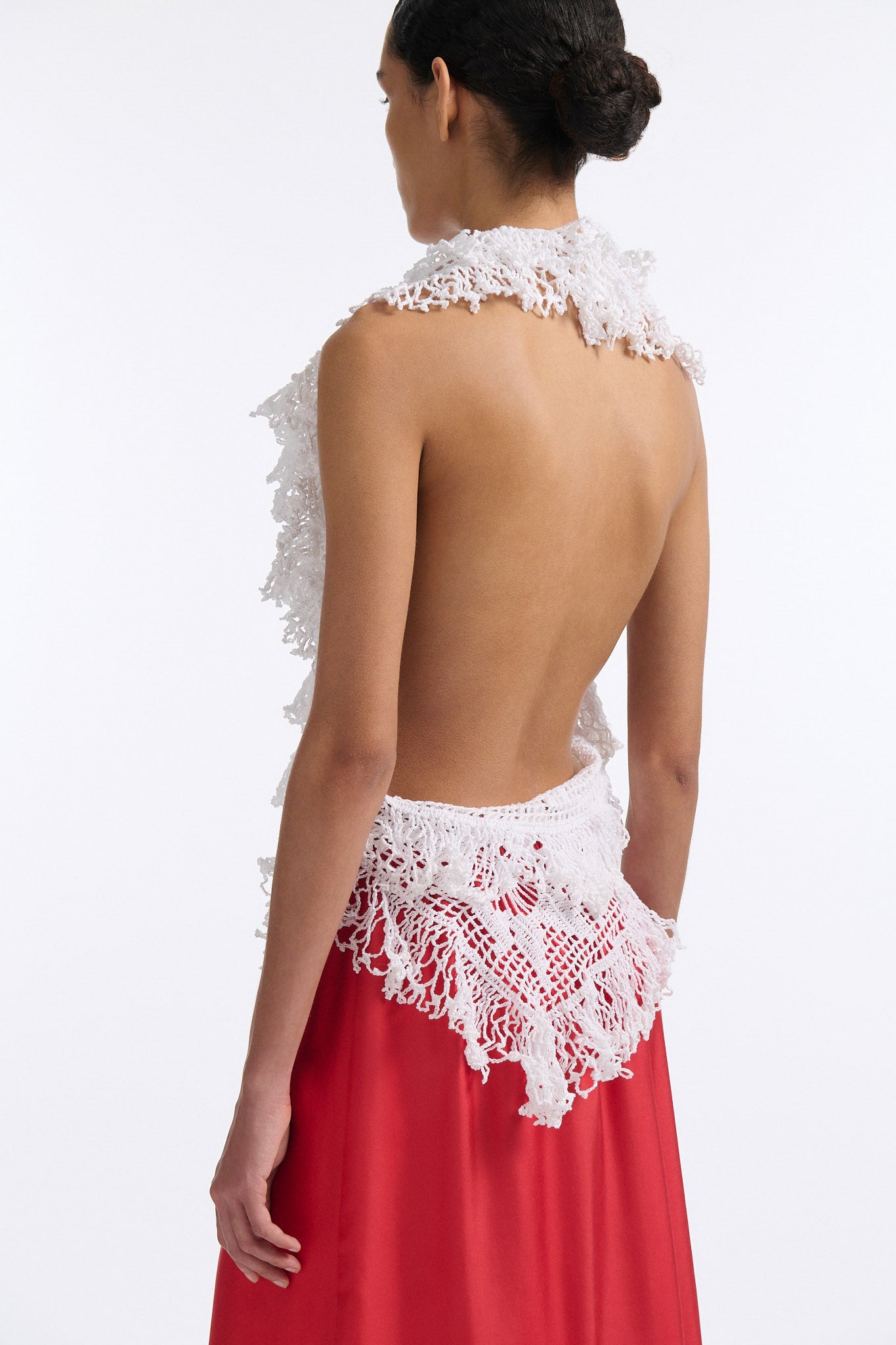 Female model with slicked back black hair in a bun wears white crochet V-neck sleeveless top with fringe crochet finishings and open back. Model also wears red silk shift skirt with cutout palm leaf hem and elastic waist.