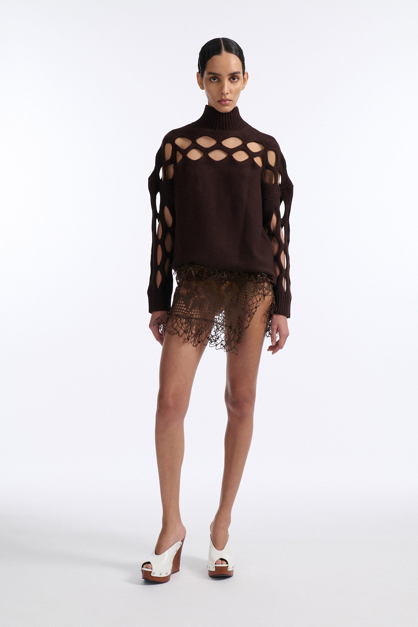 Female model with slicked back black hair in a bun wears brown knit mockneck with horizontal diamond cutouts along arms, chest, and back. Model also wears a see-through crochet mini skirt in brown and white wedges.