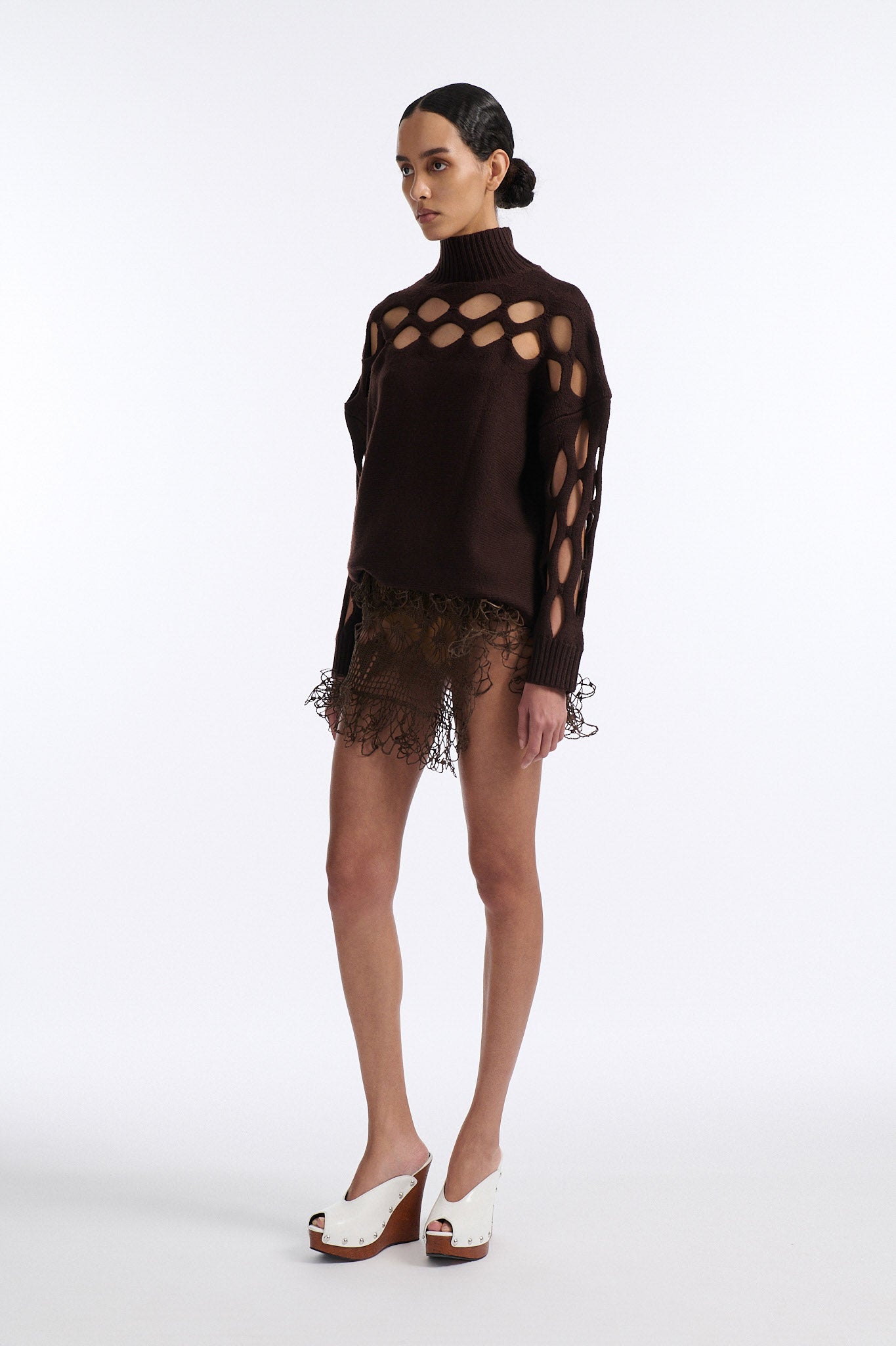Female model with slicked back black hair in a bun wears brown knit mockneck with horizontal diamond cutouts along arms, chest, and back. Model also wears a see-through crochet mini skirt in brown and white wedges.
