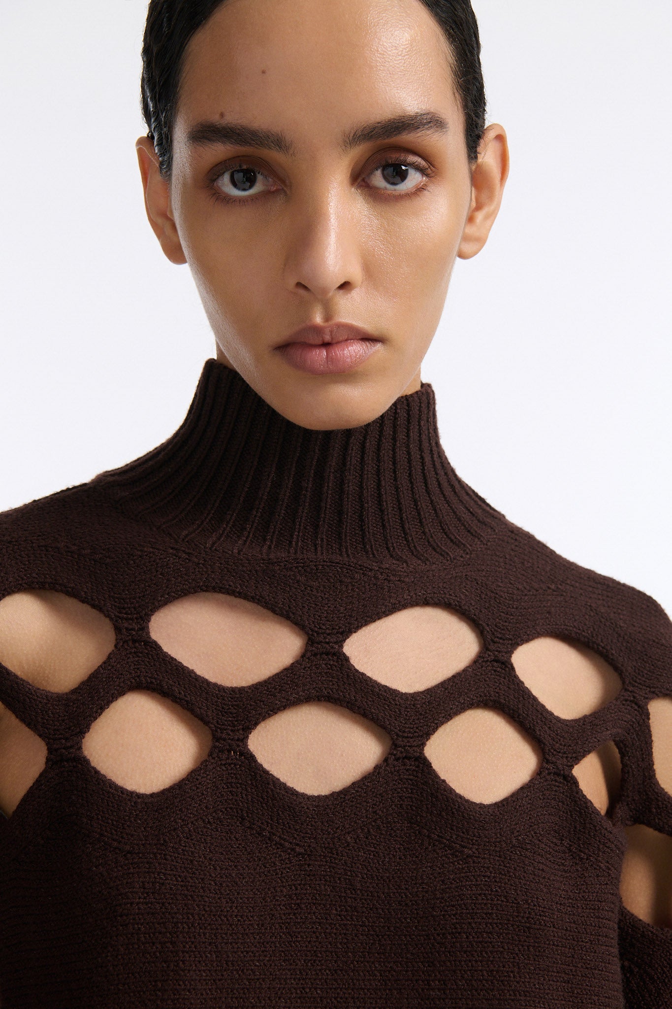 Female model with slicked back black hair in a bun wears brown knit mockneck with horizontal diamond cutouts along arms, chest, and back. Model also wears a see-through crochet mini skirt in brown and white wedges.