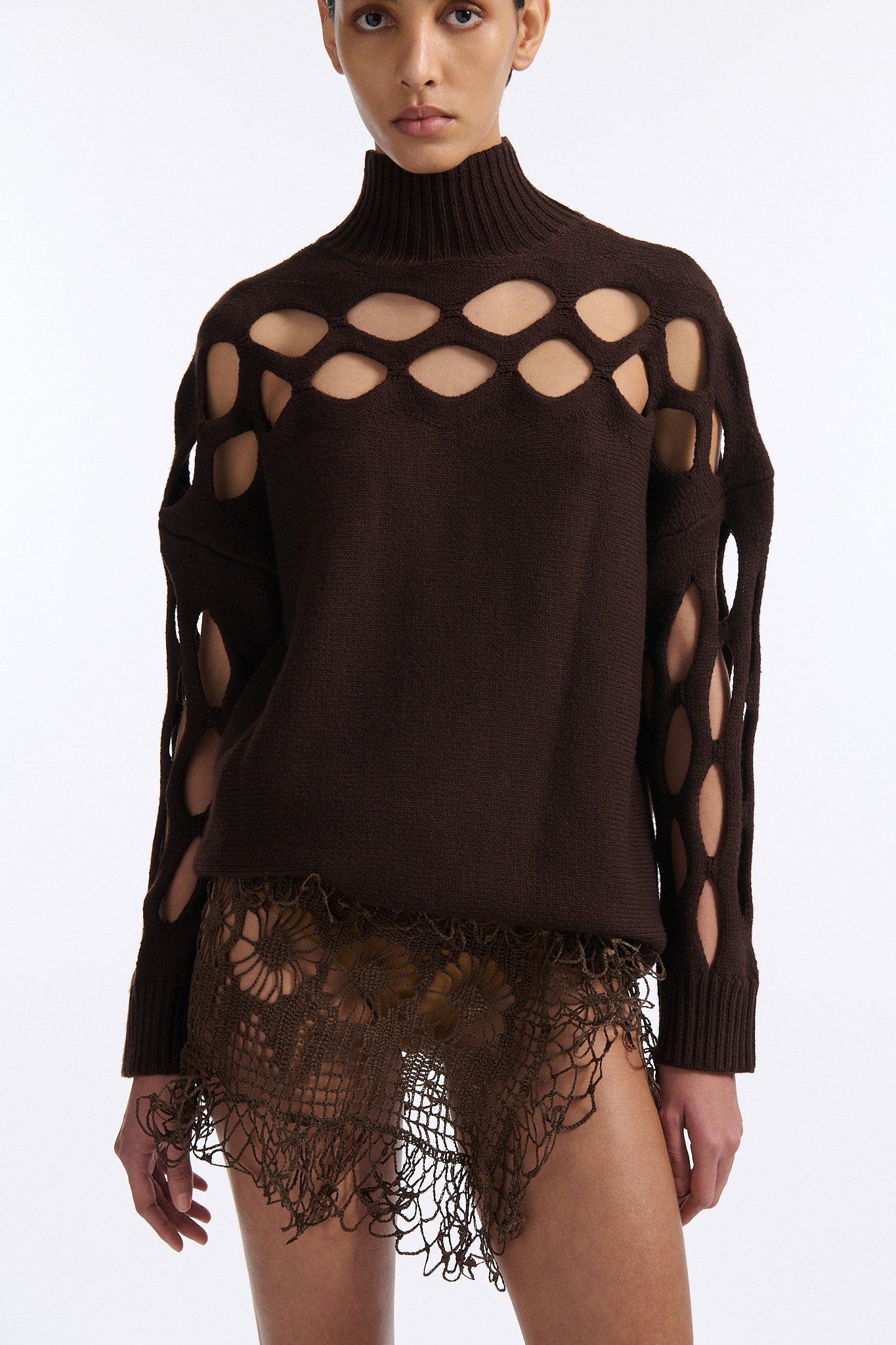 Female model with slicked back black hair in a bun wears brown knit mockneck with horizontal diamond cutouts along arms, chest, and back. Model also wears a see-through crochet mini skirt in brown and white wedges.